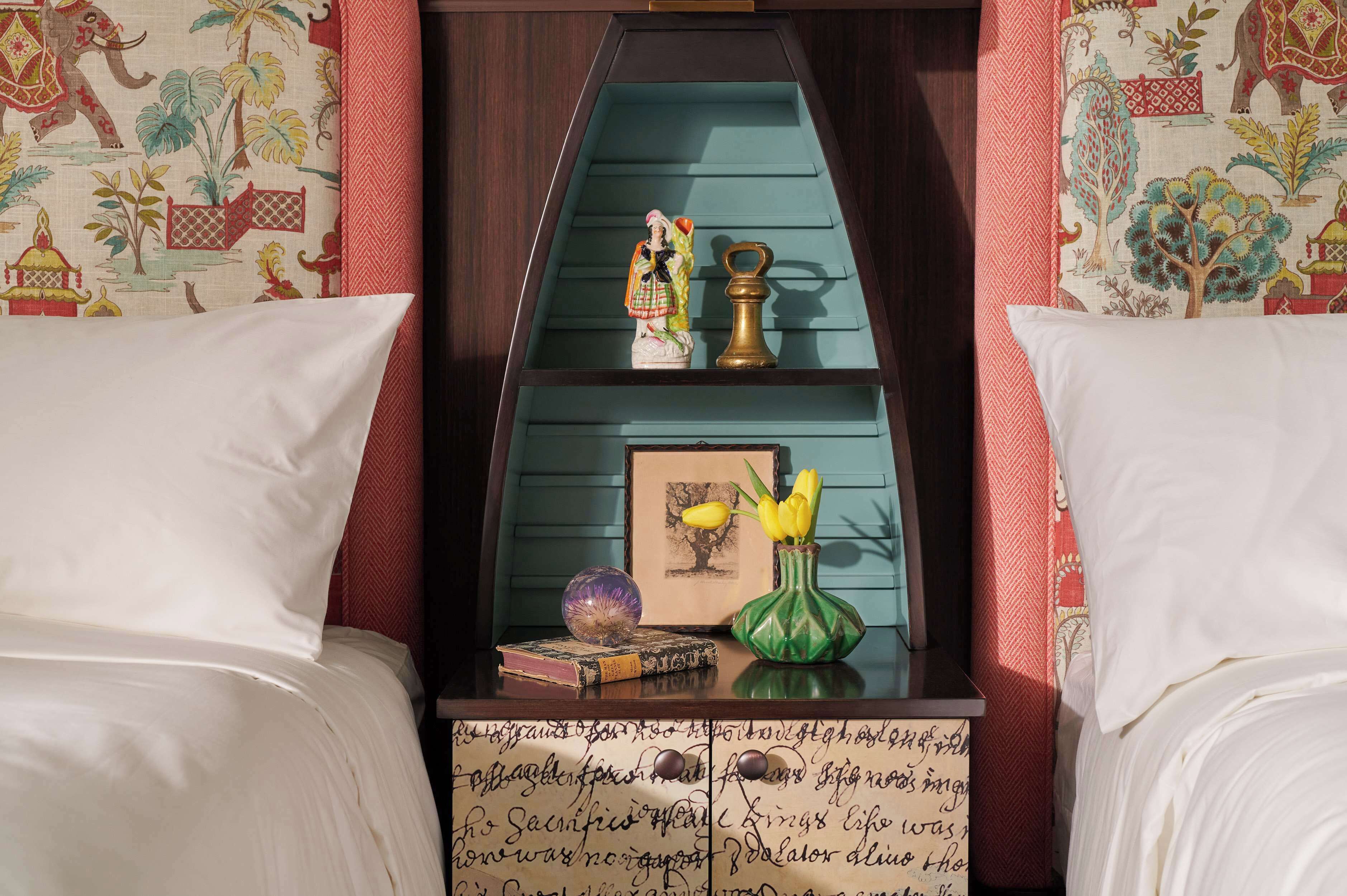 A bedroom with a bed, a nightstand, a shelf with decorative items, and a wall with patterned wallpaper.
