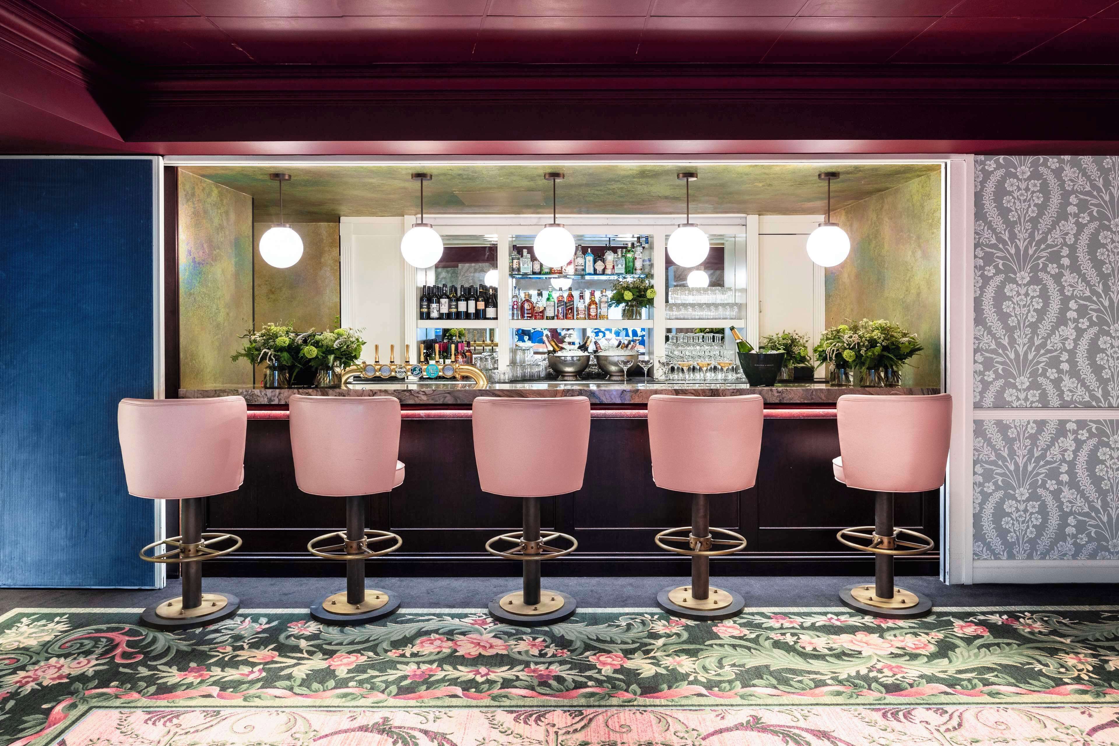 A bar area with four pink bar stools, a counter with various bottles and glasses, and hanging lights.