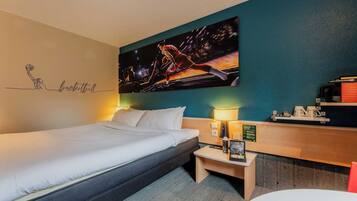 A hotel room with a bed, a desk with a lamp, a coffee machine, and a painting of a basketball game on the wall.