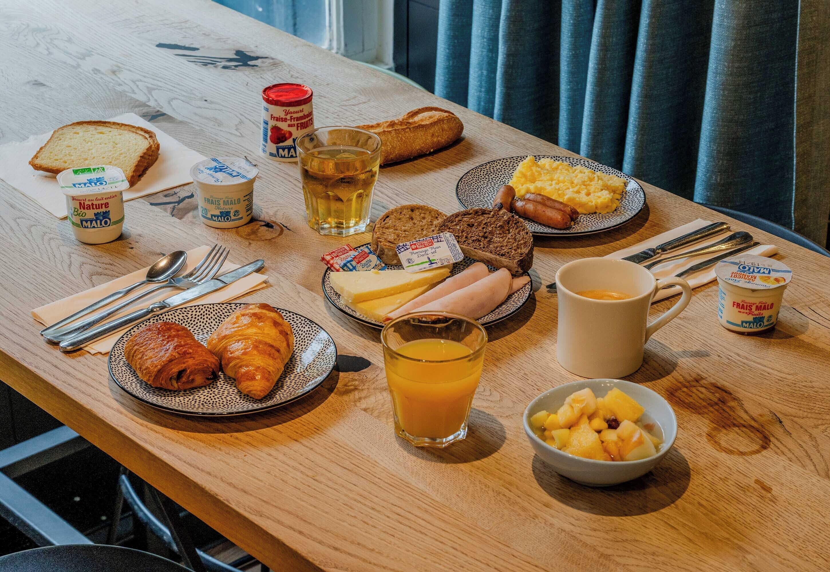 A breakfast table with various food items including bread, croissants, cheese, ham, eggs, sausages, and fruit.
