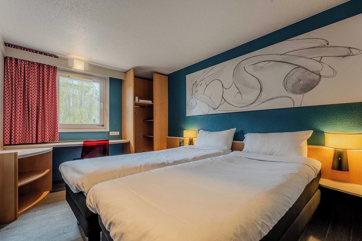 A hotel room with two beds, a desk, a chair, a window with red curtains, and a wall art piece.