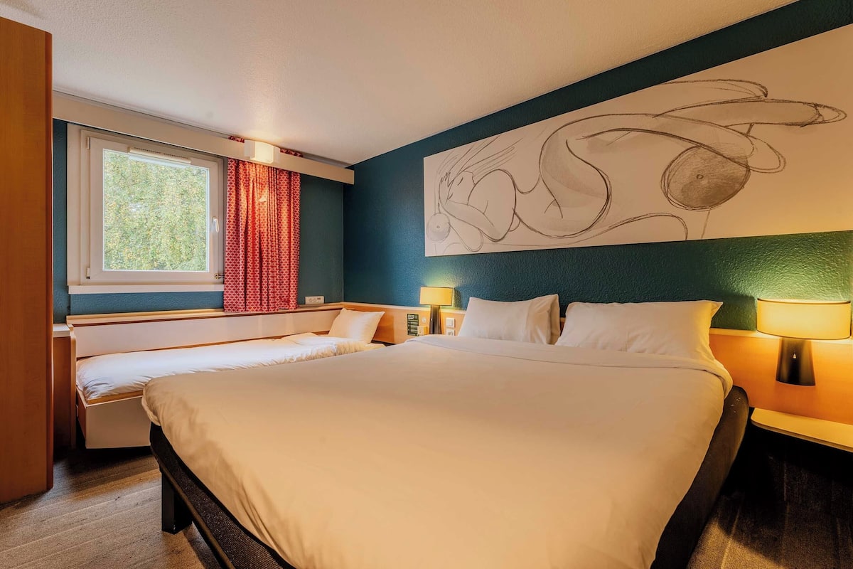 A hotel room with a large bed, a smaller bed, two bedside tables with lamps, a window with red curtains, and a wall mural.