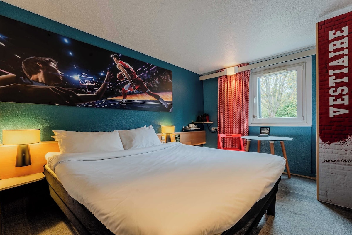 A hotel room with a bed, a nightstand, a wall-mounted TV, a window with curtains, and a red wall with the word "VESTIARE" on it.