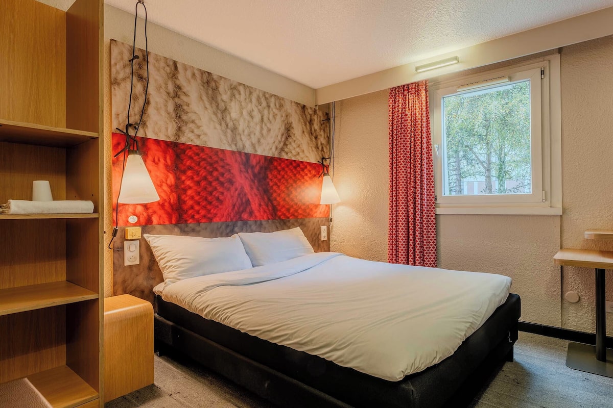 A hotel room with a bed, bedside lamps, a window with curtains, and a headboard with a patterned design.