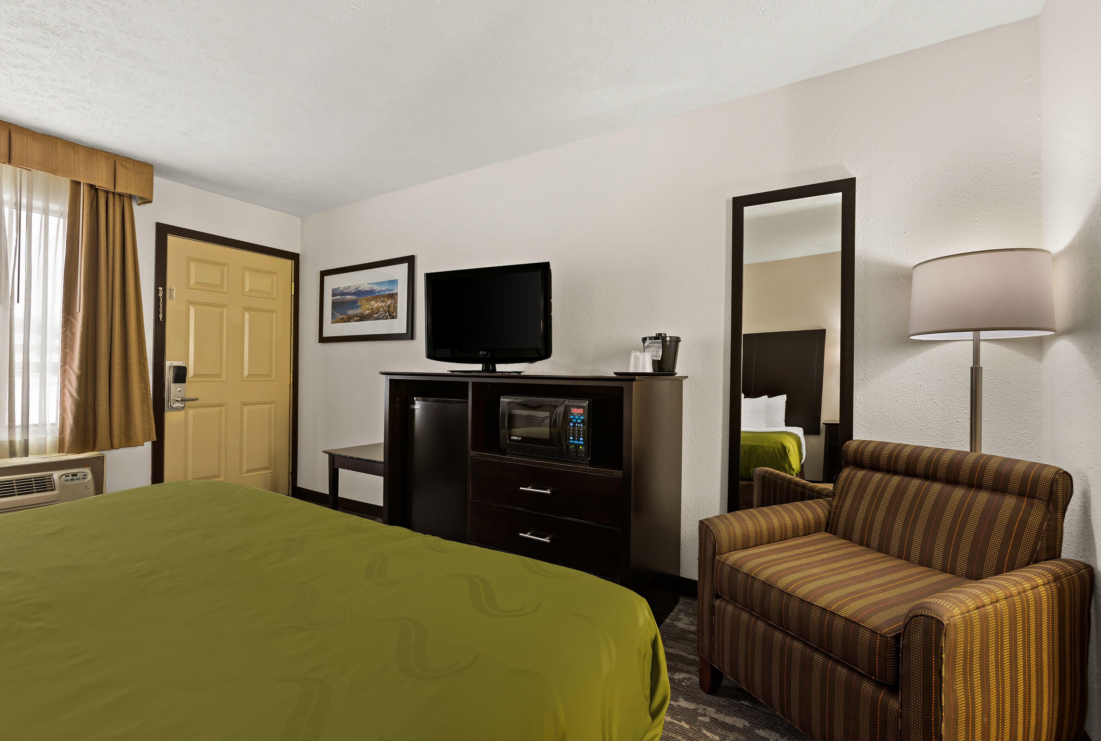 A hotel room with a bed, a desk with a television, a microwave, a chair, and a lamp.
