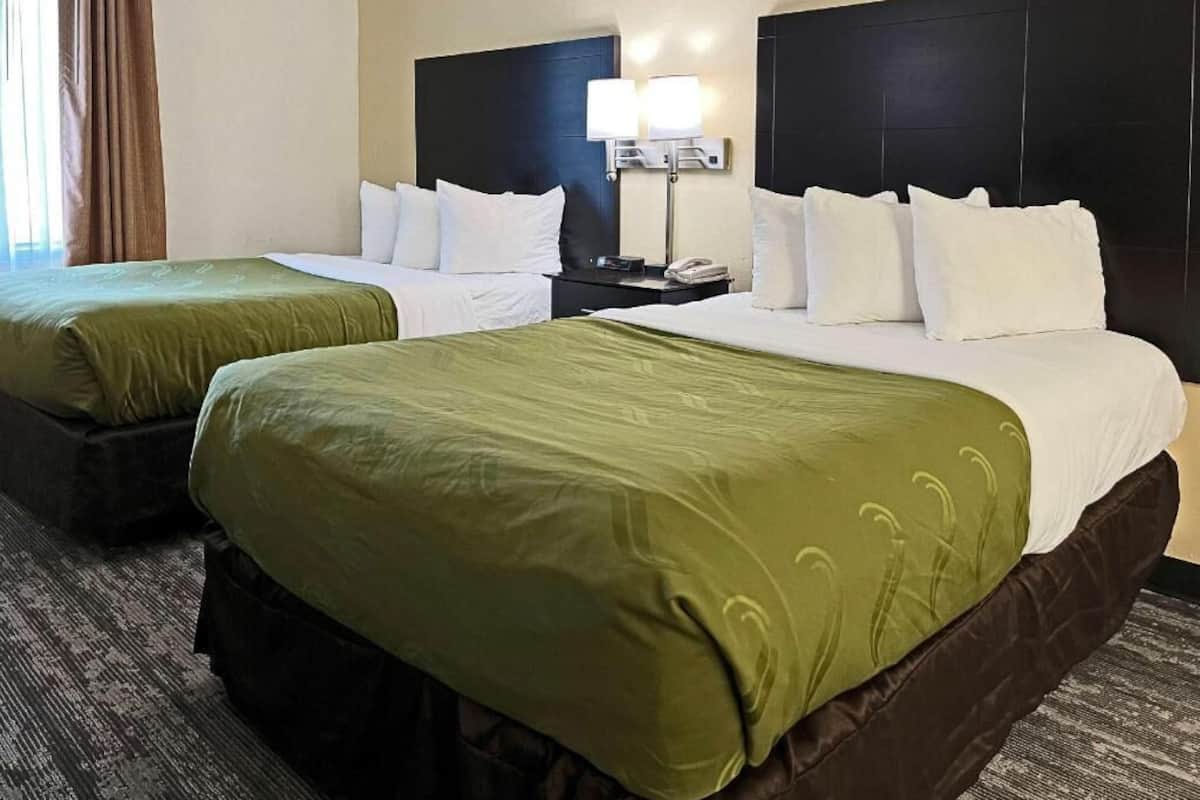 A hotel room with two beds, a nightstand, a lamp, and a window with curtains.