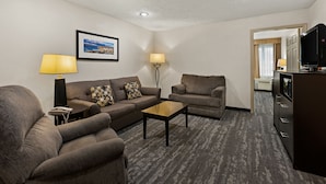 Une chambre d’hôtel avec un canapé, un fauteuil et une petite table. On y trouve également une télévision et une lampe sur un support.