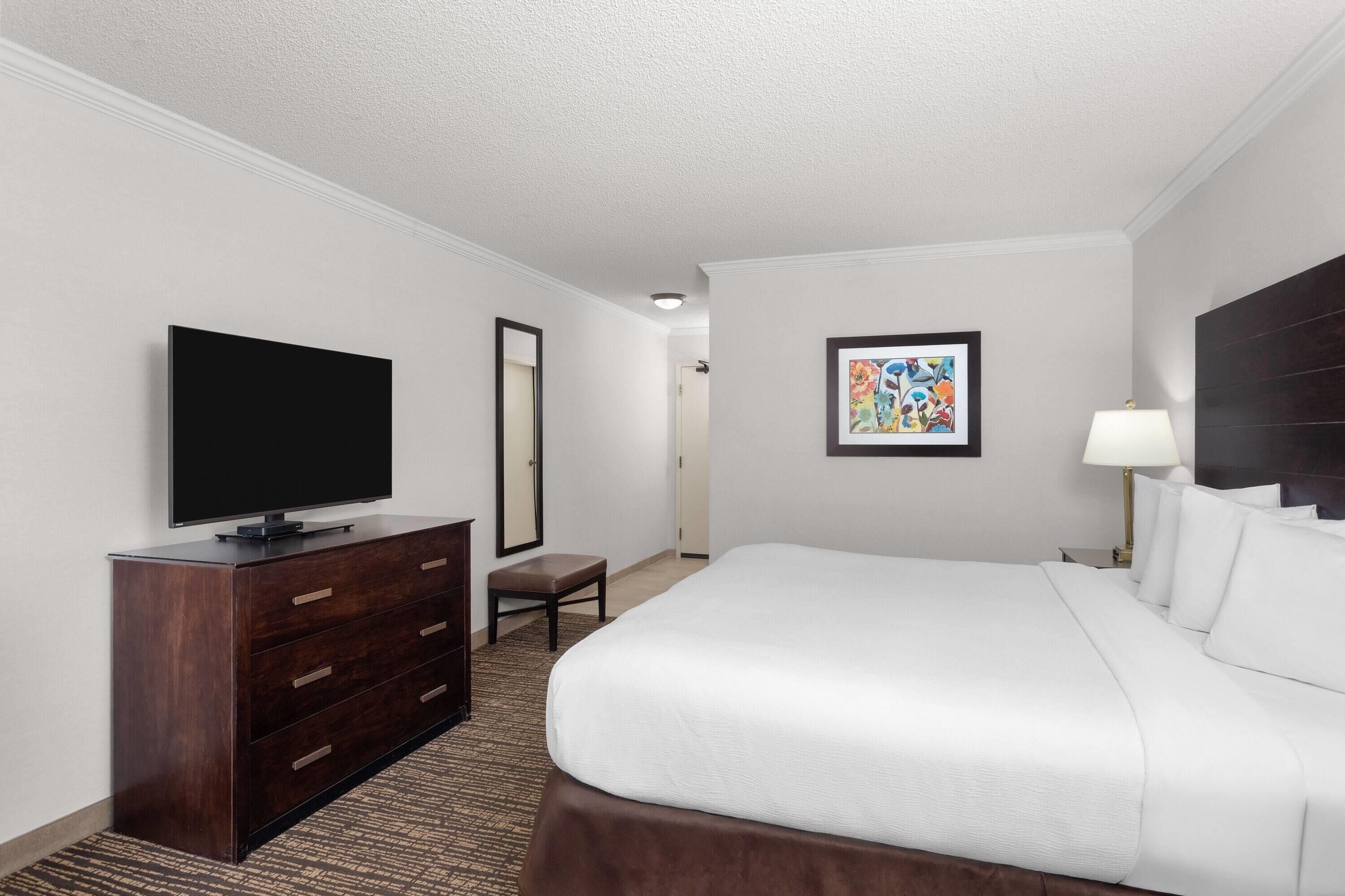 A hotel room with a large bed, a flat-screen TV on a wooden dresser, a small table, and a framed artwork on the wall.