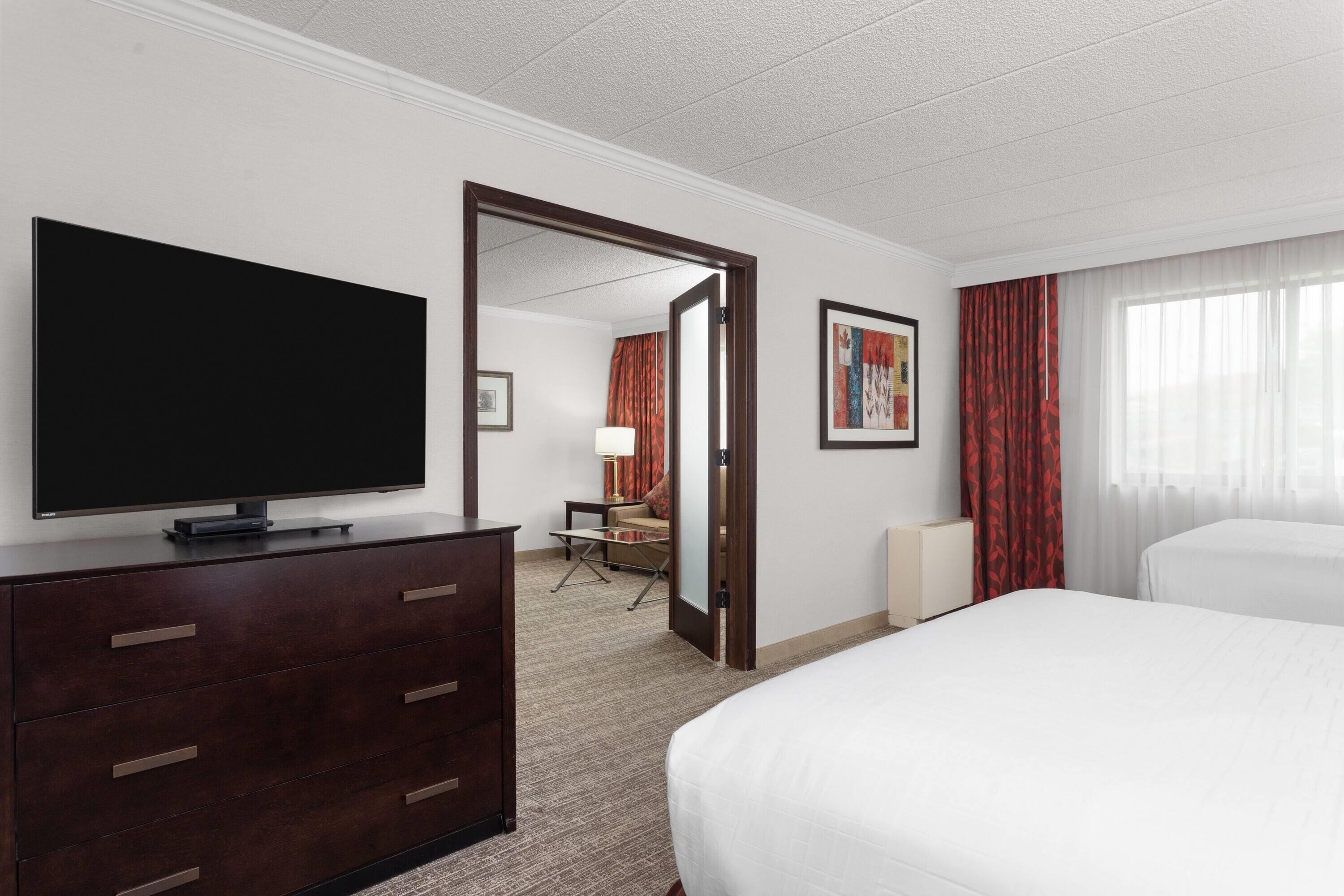 A hotel room with a large flat-screen TV on a dresser, a bed, a desk with a chair, and a door leading to another room.
