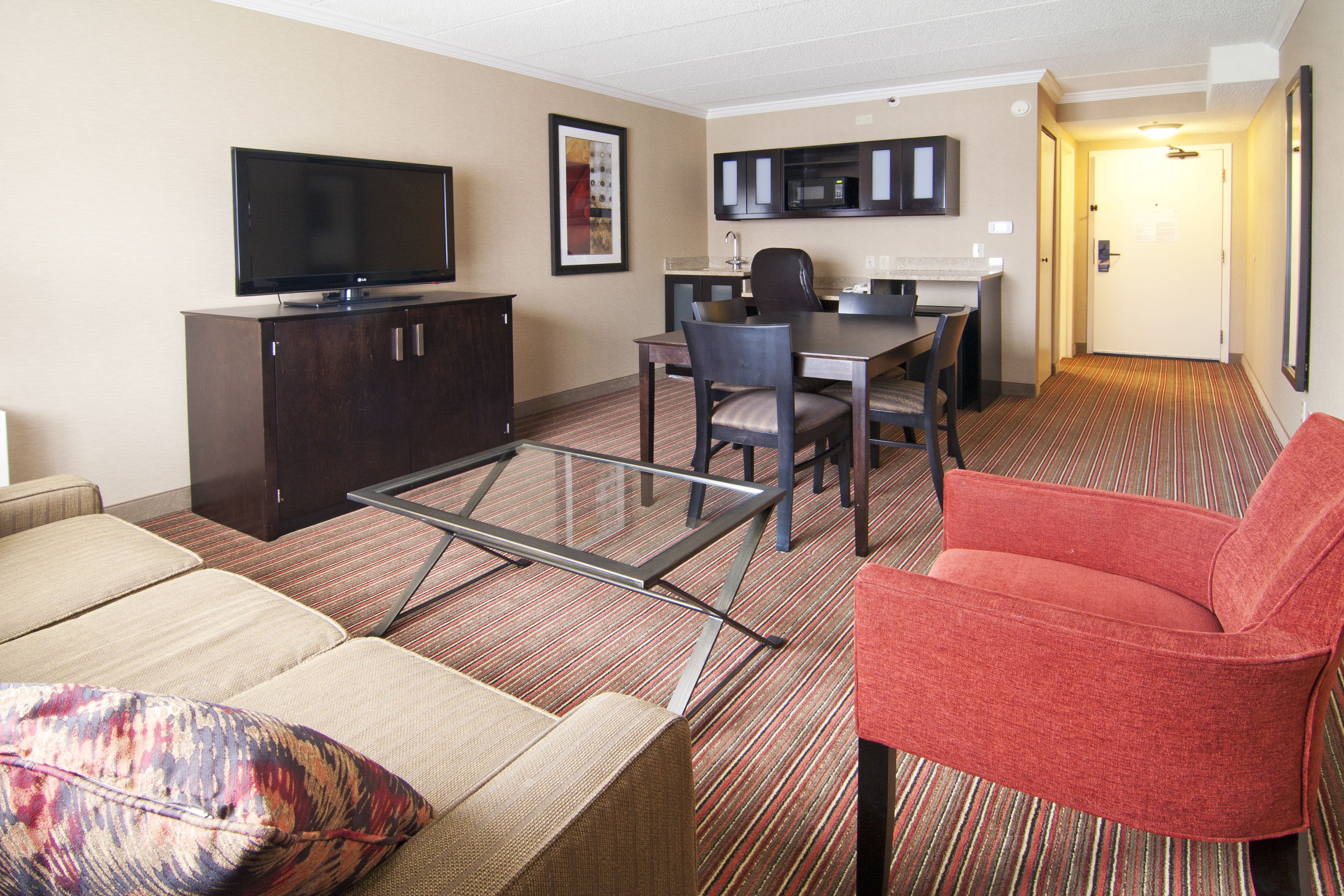 A hotel room with a flat-screen TV, a dining table with chairs, a red armchair, a sofa, and a carpeted floor.