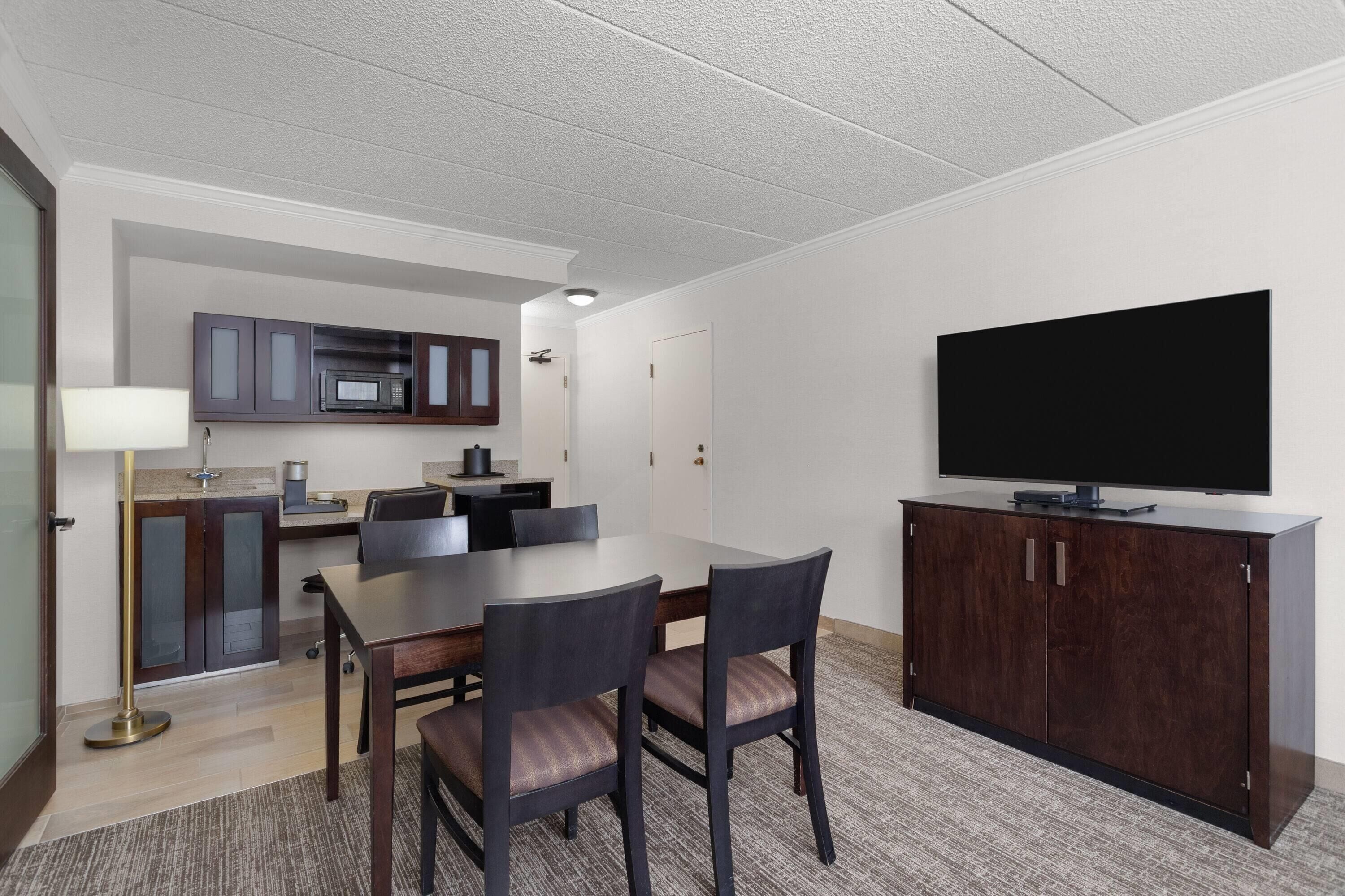 A modern hotel room with a dining area, a flat-screen TV, and a kitchenette.