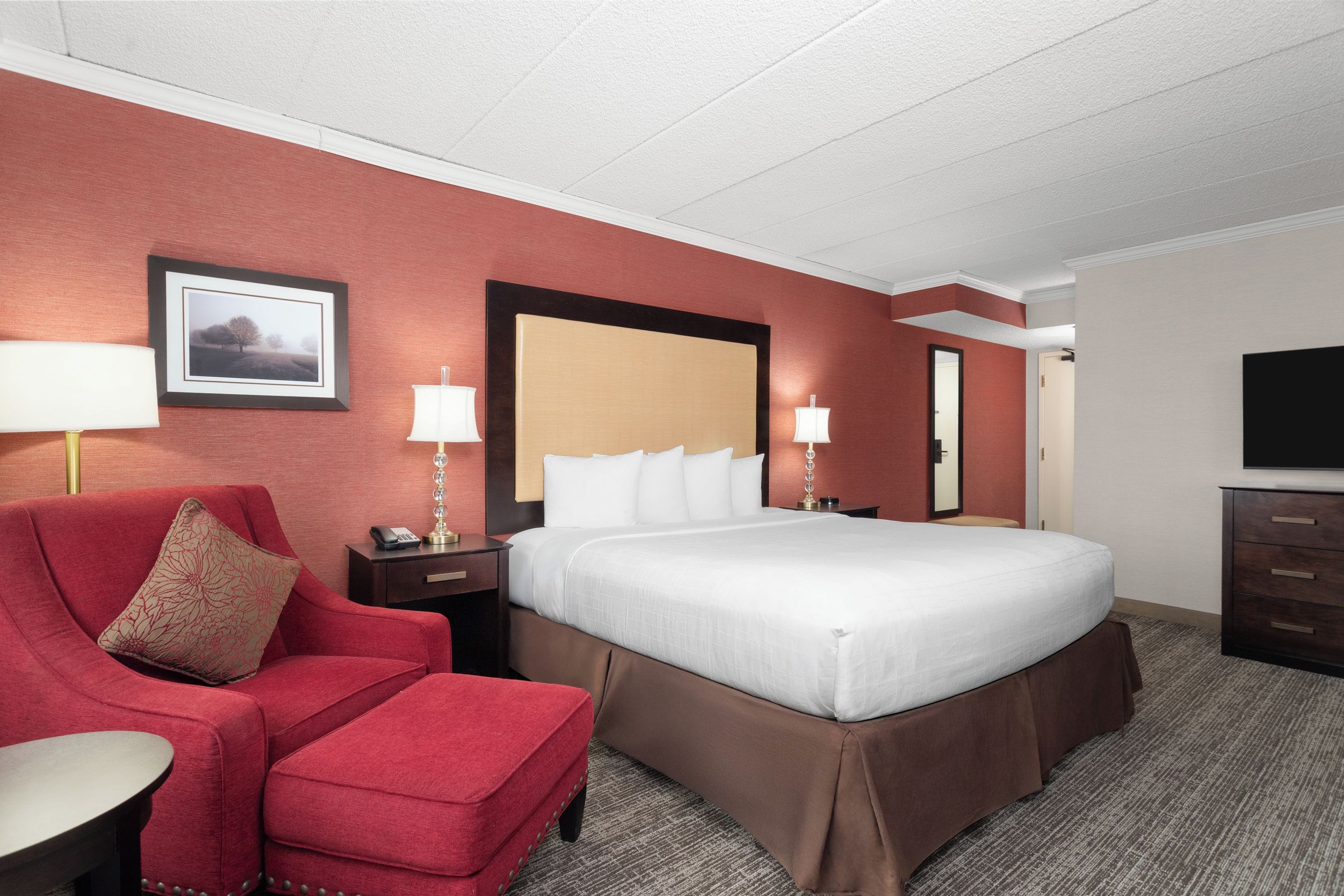 A hotel room with a large bed, a red armchair, a desk with a lamp, and a television.
