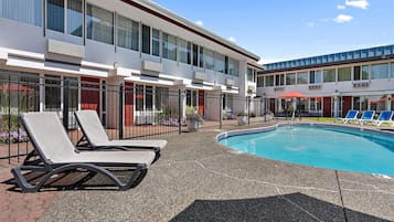 Piscina com espreguiçadeiras, um edifício com várias janelas e céu claro.