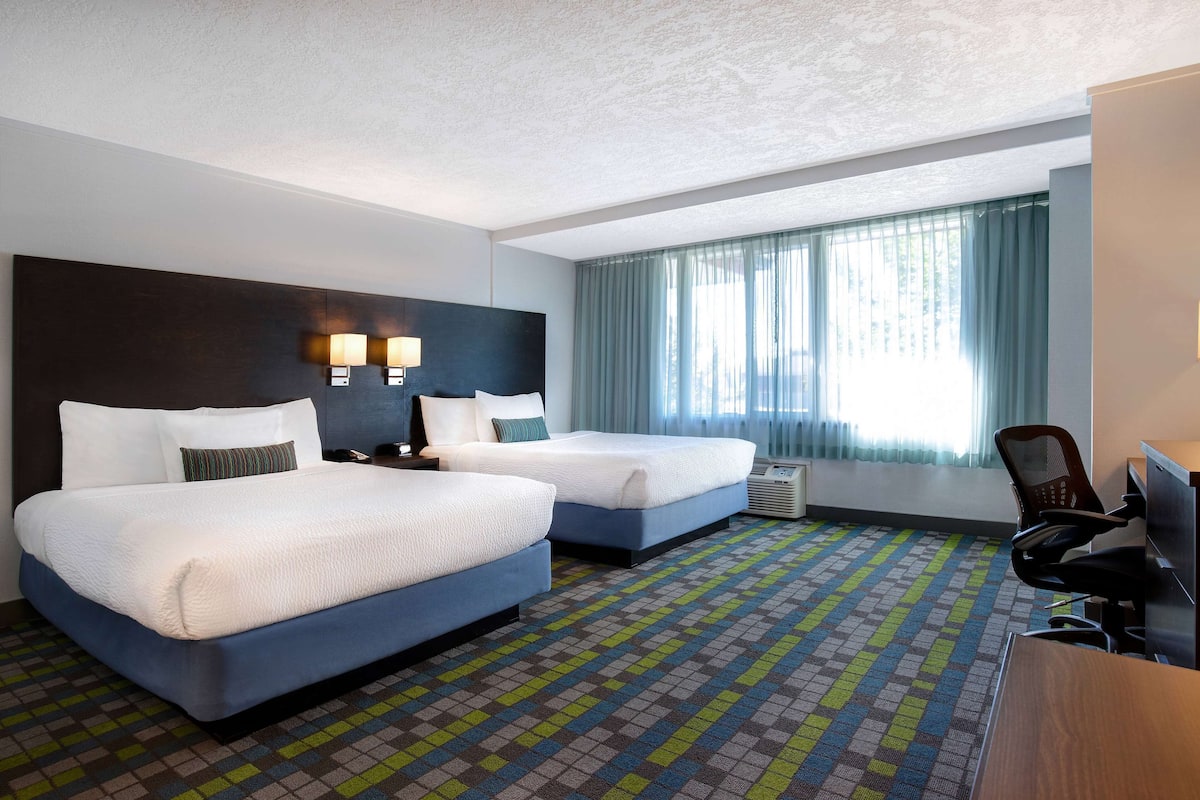 Ein Hotelzimmer mit zwei Betten, einem Schreibtisch, einem Stuhl und einem großen Fenster mit Vorhängen.