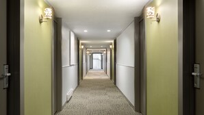 Een hotelgang met vloerbedekking, lichte wanden en wandlampen.