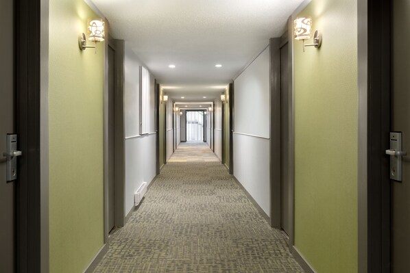 Un couloir d’hôtel avec des revêtements de sol en moquette, des murs de couleur claire et des luminaires fixés au mur.