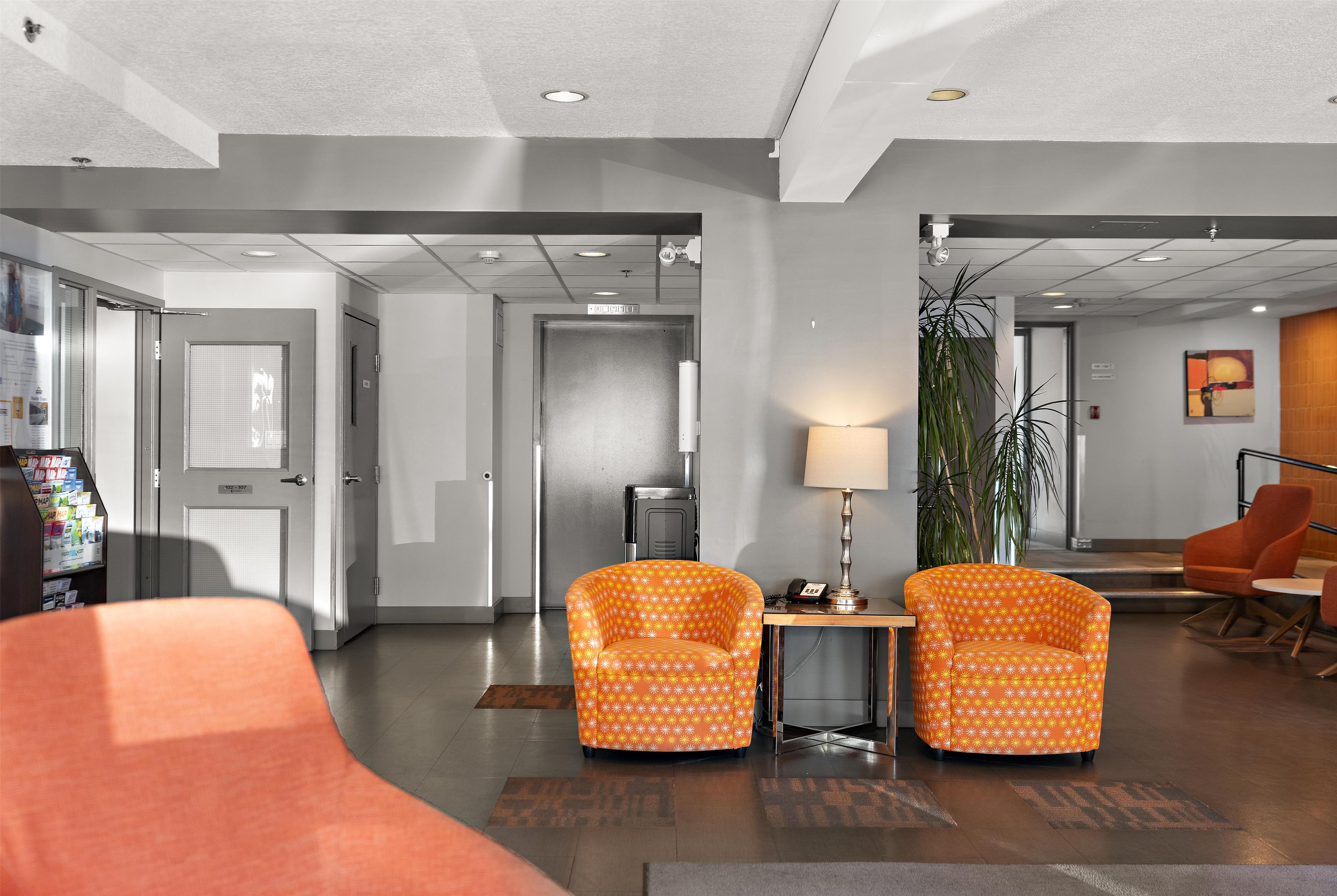A hotel lobby with a reception desk, two patterned armchairs, a coffee table, a lamp, a plant, and a large orange sofa.
