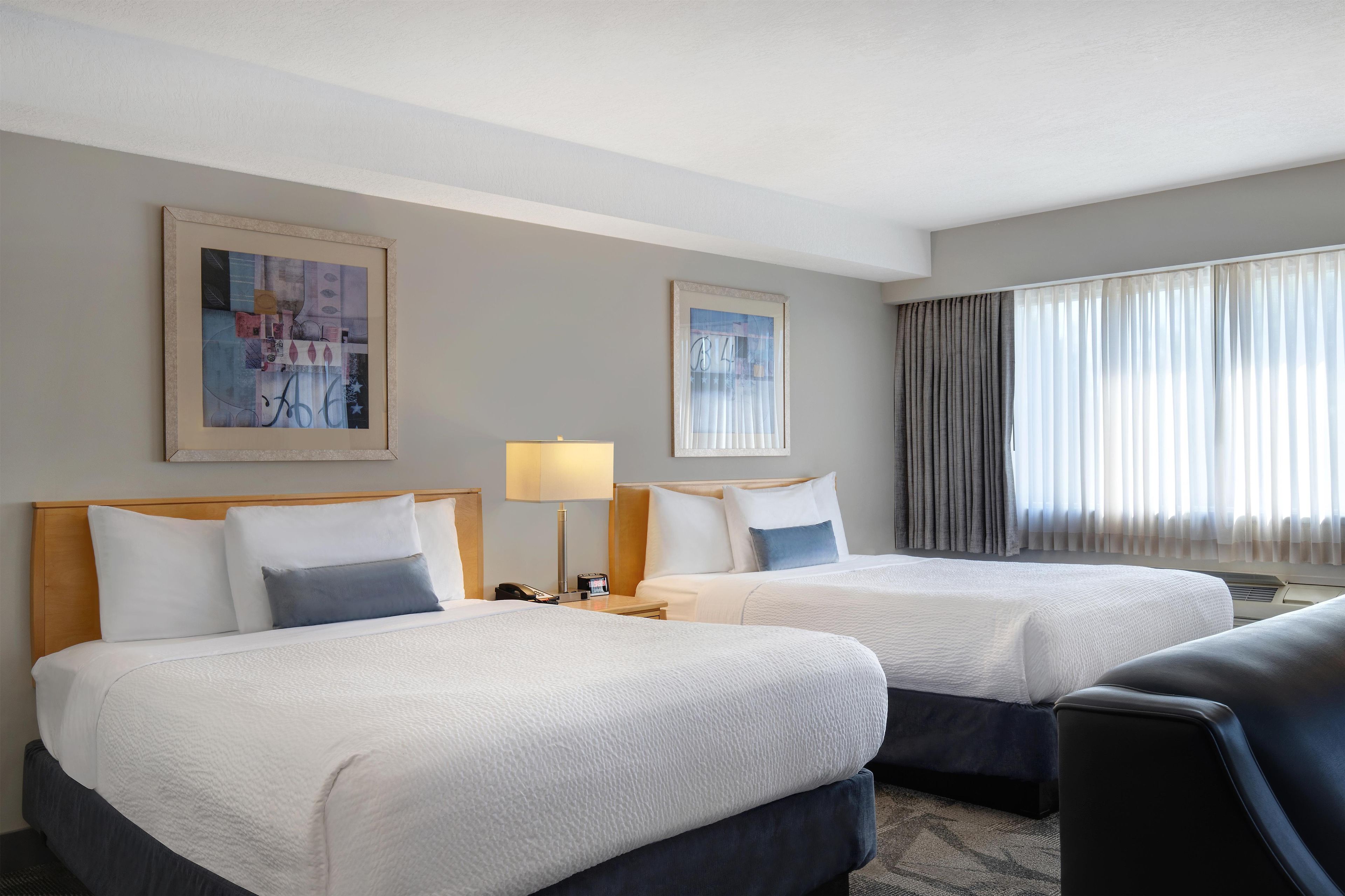 Habitación de hotel con dos camas, una mesita de noche con lámpara, una ventana con cortinas y dos cuadros en la pared.