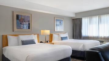 Habitación de hotel con dos camas, una mesita de noche con lámpara, una ventana con cortinas y dos cuadros en la pared.