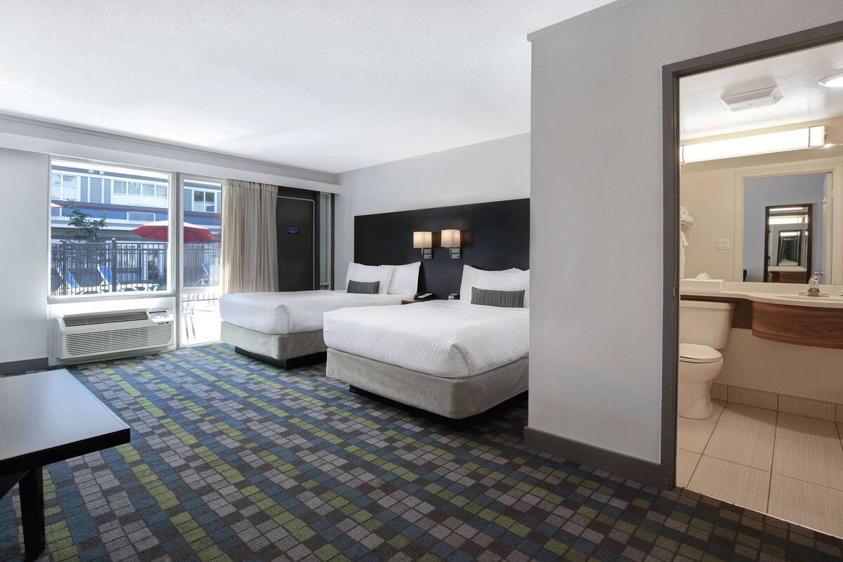 Ein Hotelzimmer mit zwei Betten, einem großen Fenster, einem Balkon, einem kleinen Tisch und einem Badezimmer mit Spiegel und Toilette.