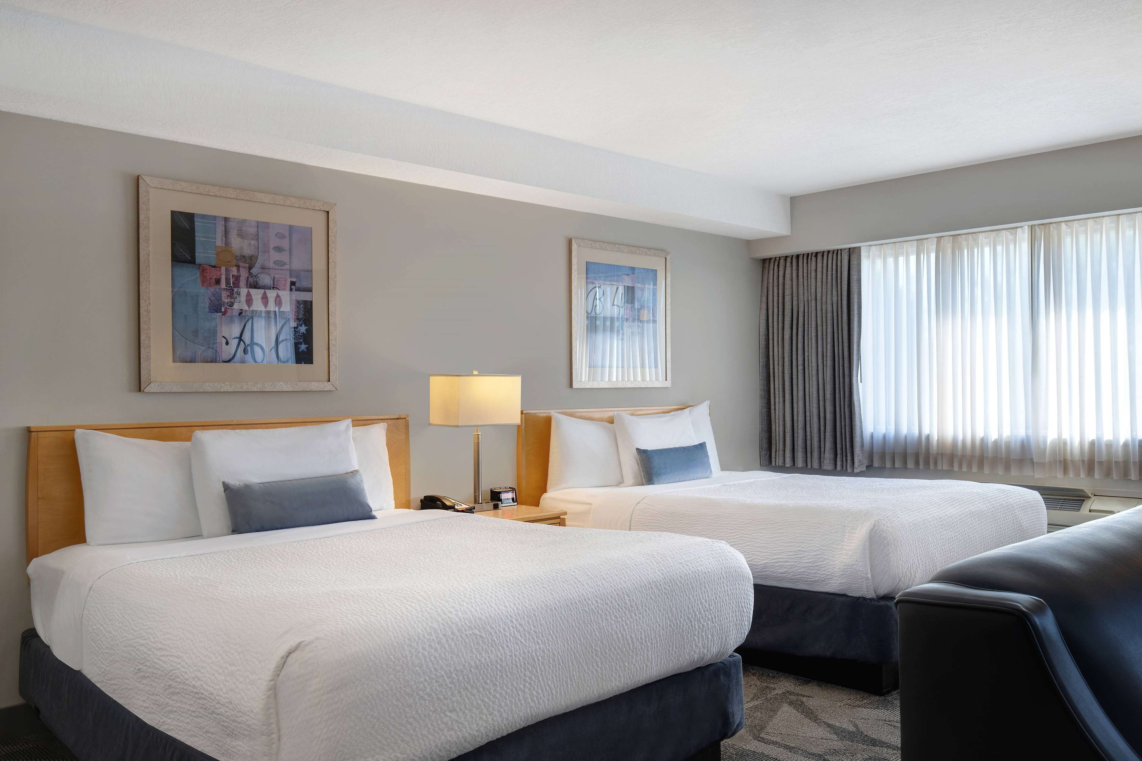 Una habitación de hotel con dos camas, una mesita de noche con lámpara y un amplio ventanal con cortinas.