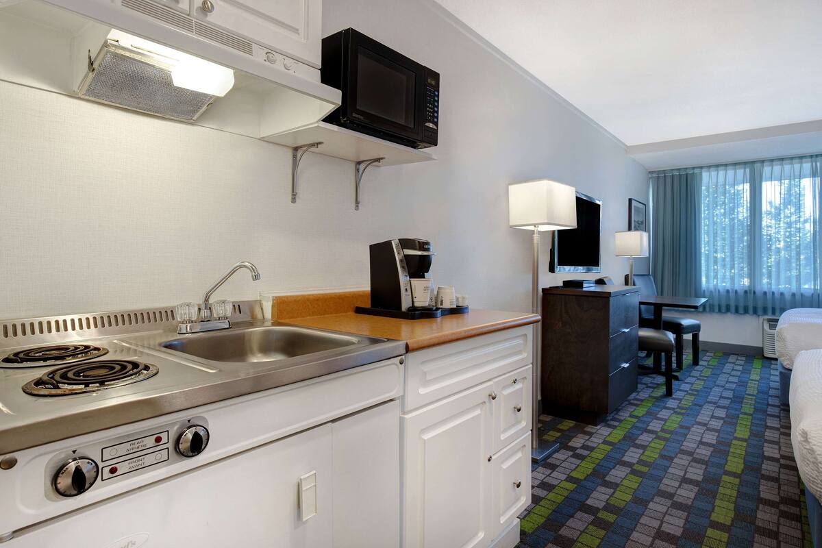 Ein Hotelzimmer mit einer Küchenzeile, inklusive Herd, Spüle und Mikrowelle. Es gibt einen Schreibtisch mit Stuhl, eine Lampe und ein Bett.