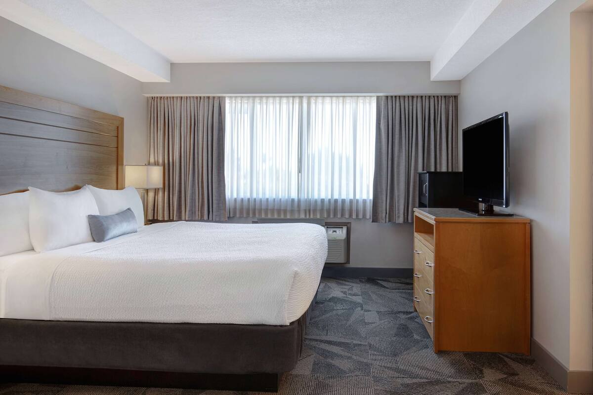 Ein Hotelzimmer mit einem großen Bett, einem Fernseher auf einer Holzkommode und einem Fenster mit Vorhängen.