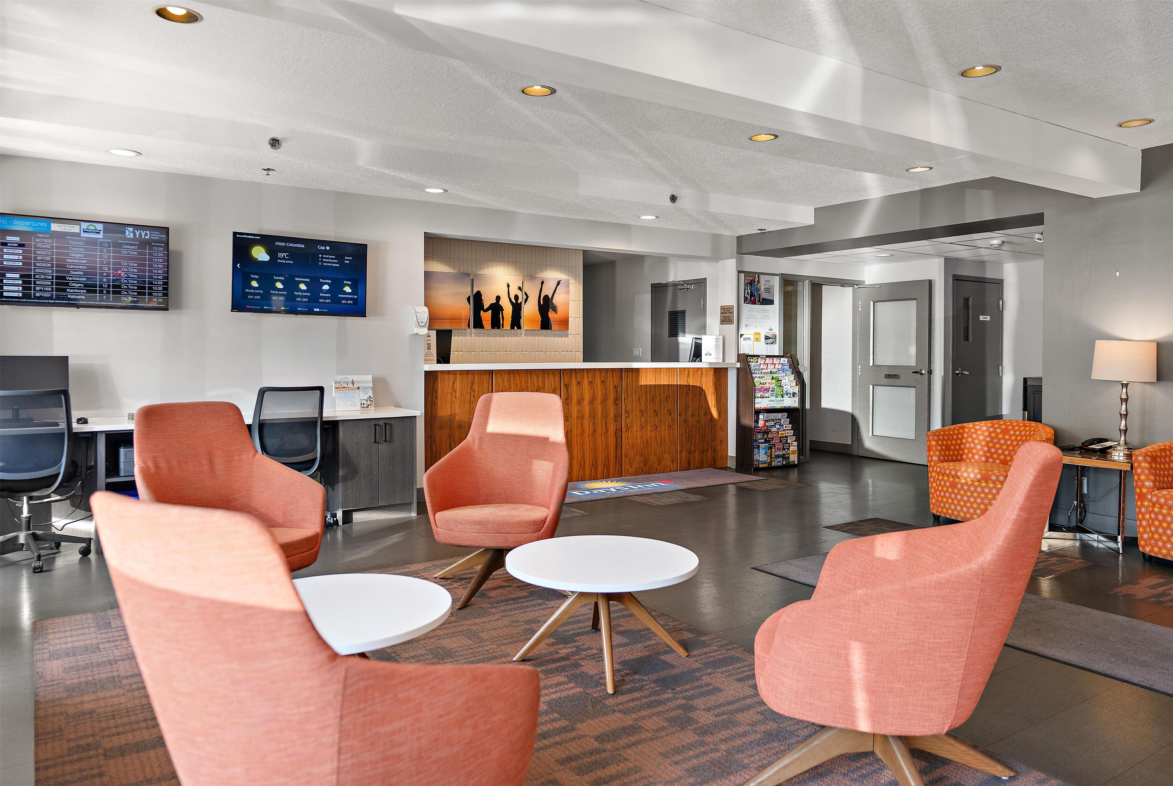 A hotel lobby with a reception desk, seating area with chairs, a television displaying flight information, and a wall-mounted artwork.