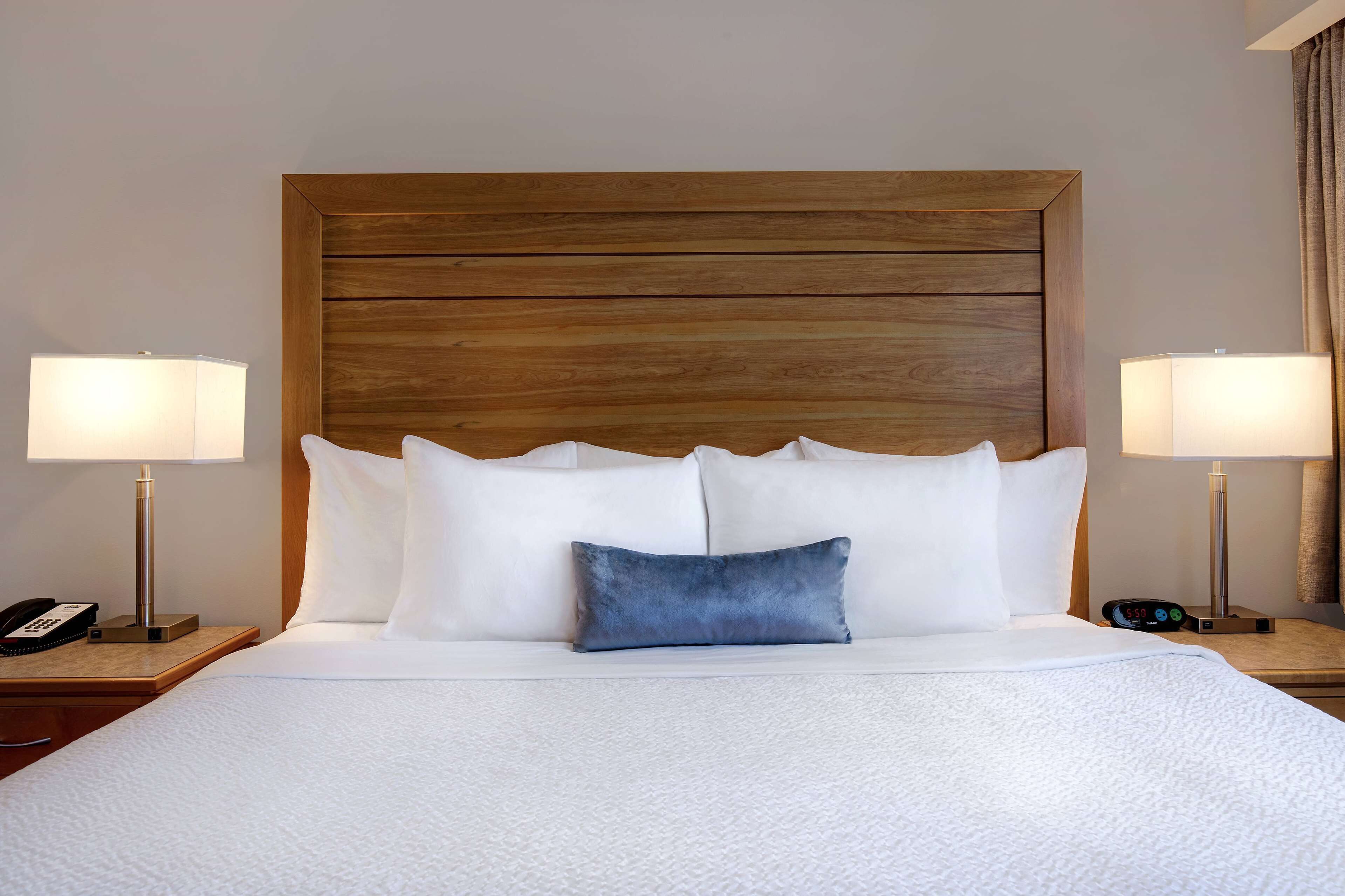 A neatly made bed with white bedding and a blue accent pillow, flanked by two bedside tables with lamps and clocks.