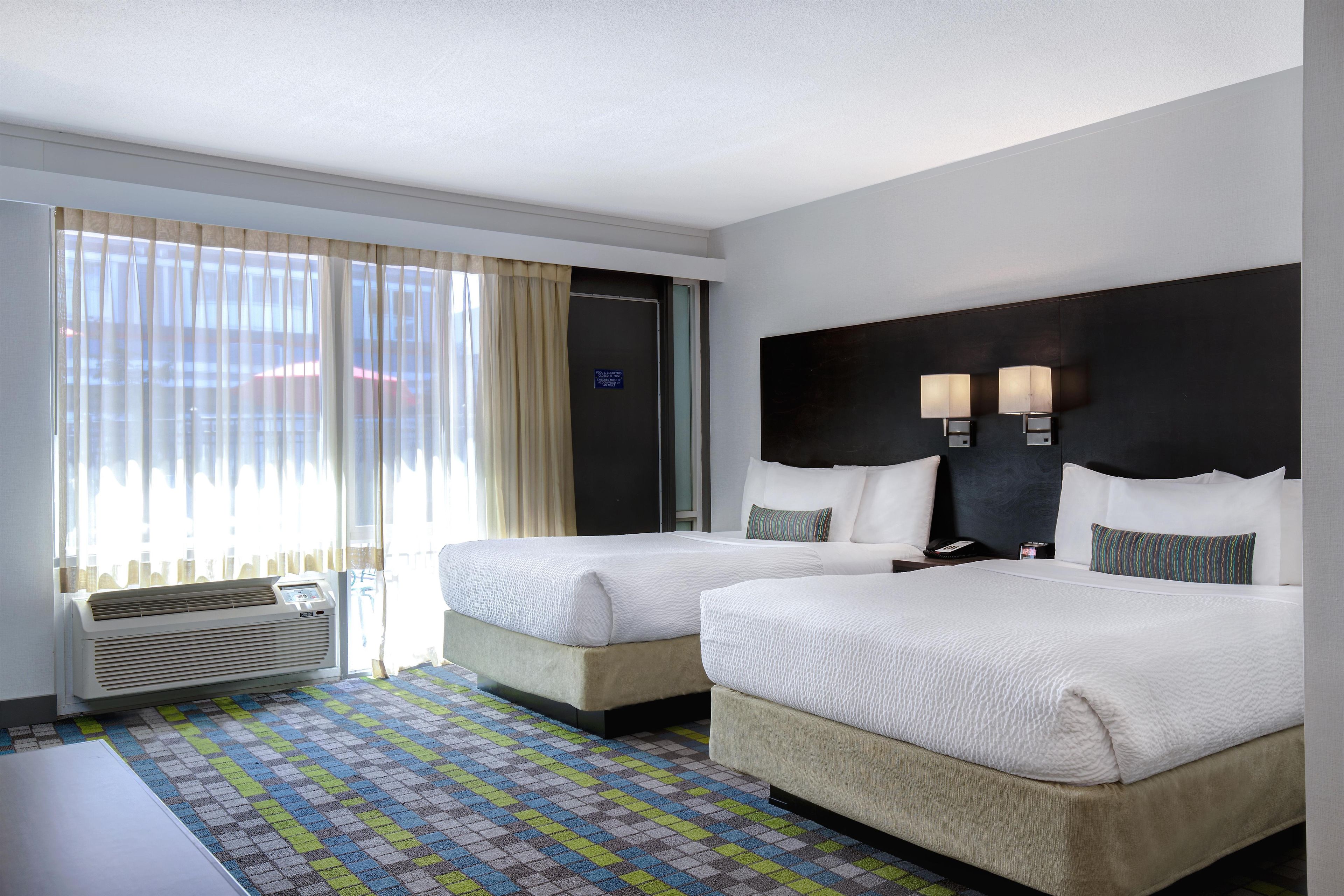 A hotel room with two beds, a large window with curtains, a wall-mounted air conditioner, and a patterned carpet.