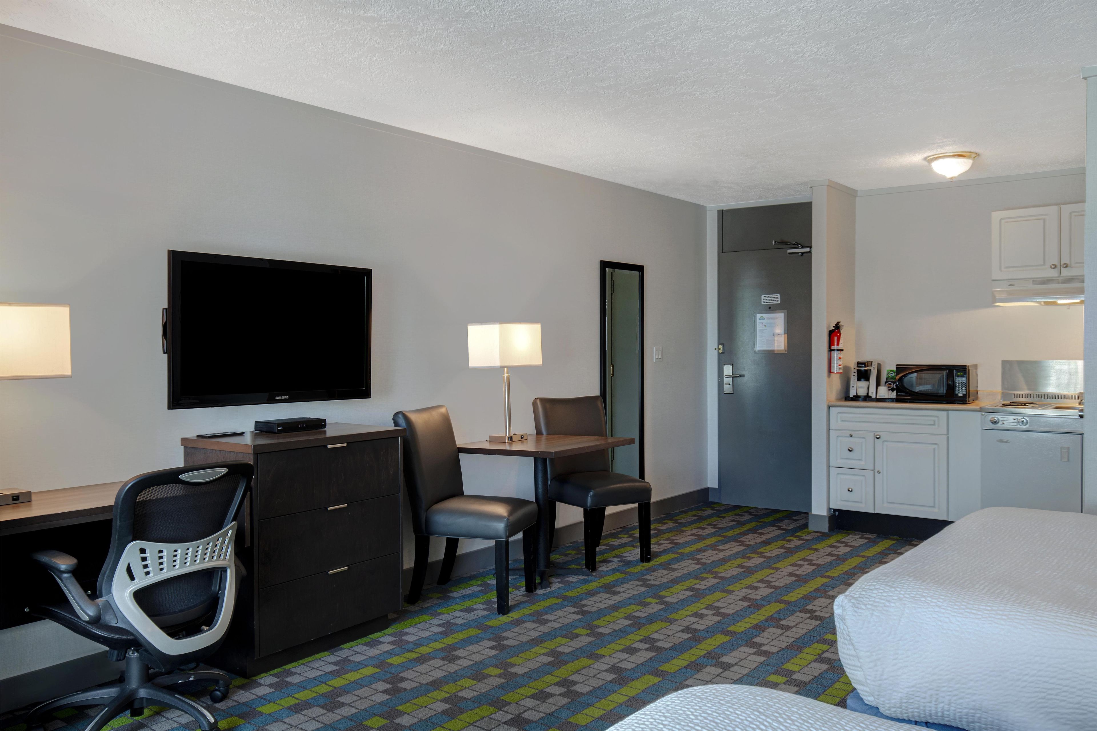 Habitación de hotel con escritorio, sillas, televisor, kitchenette y cama.