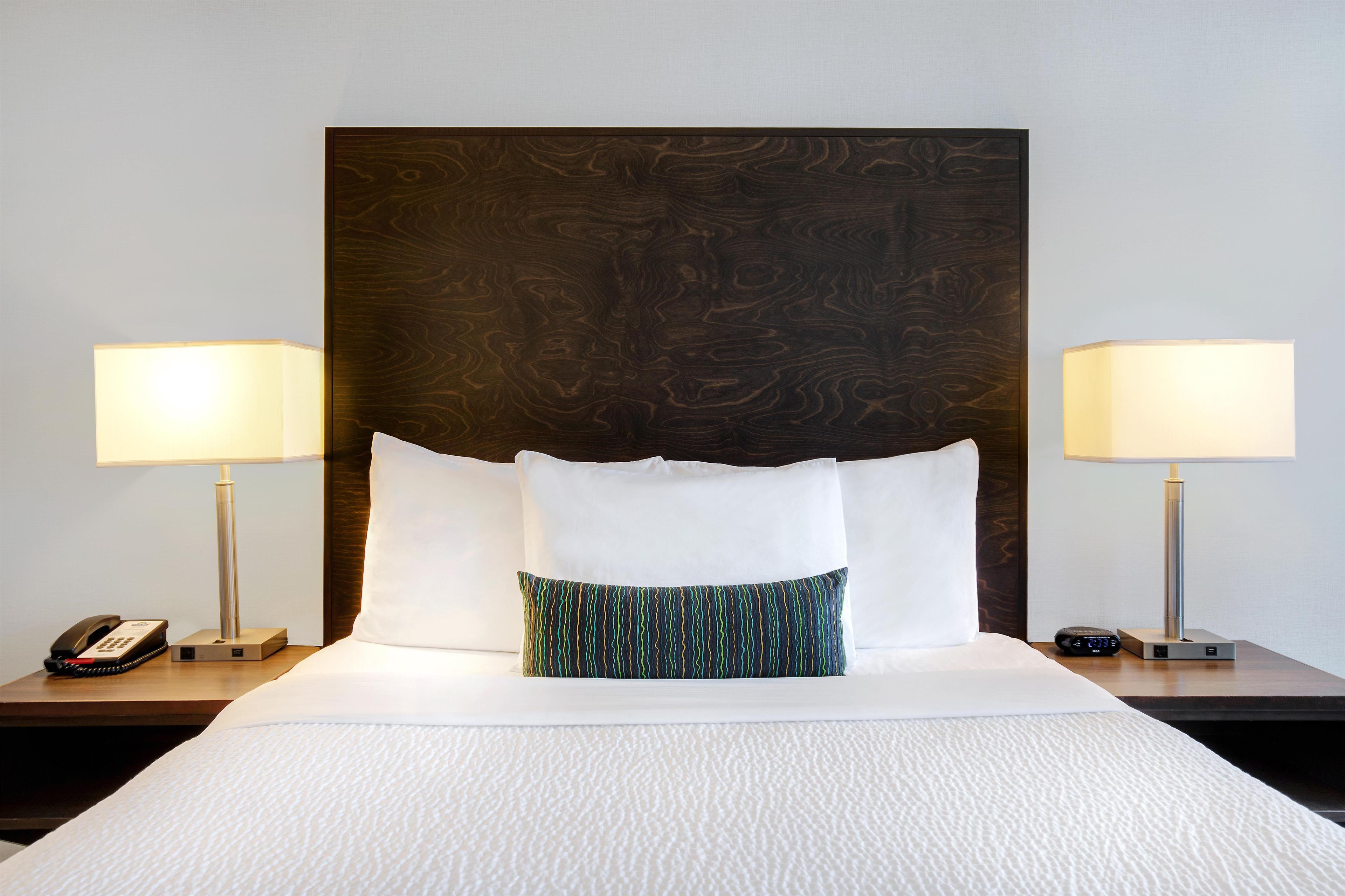 Habitación de hotel con una cama grande, dos lámparas de noche, un teléfono y un reloj digital en la mesita de noche.