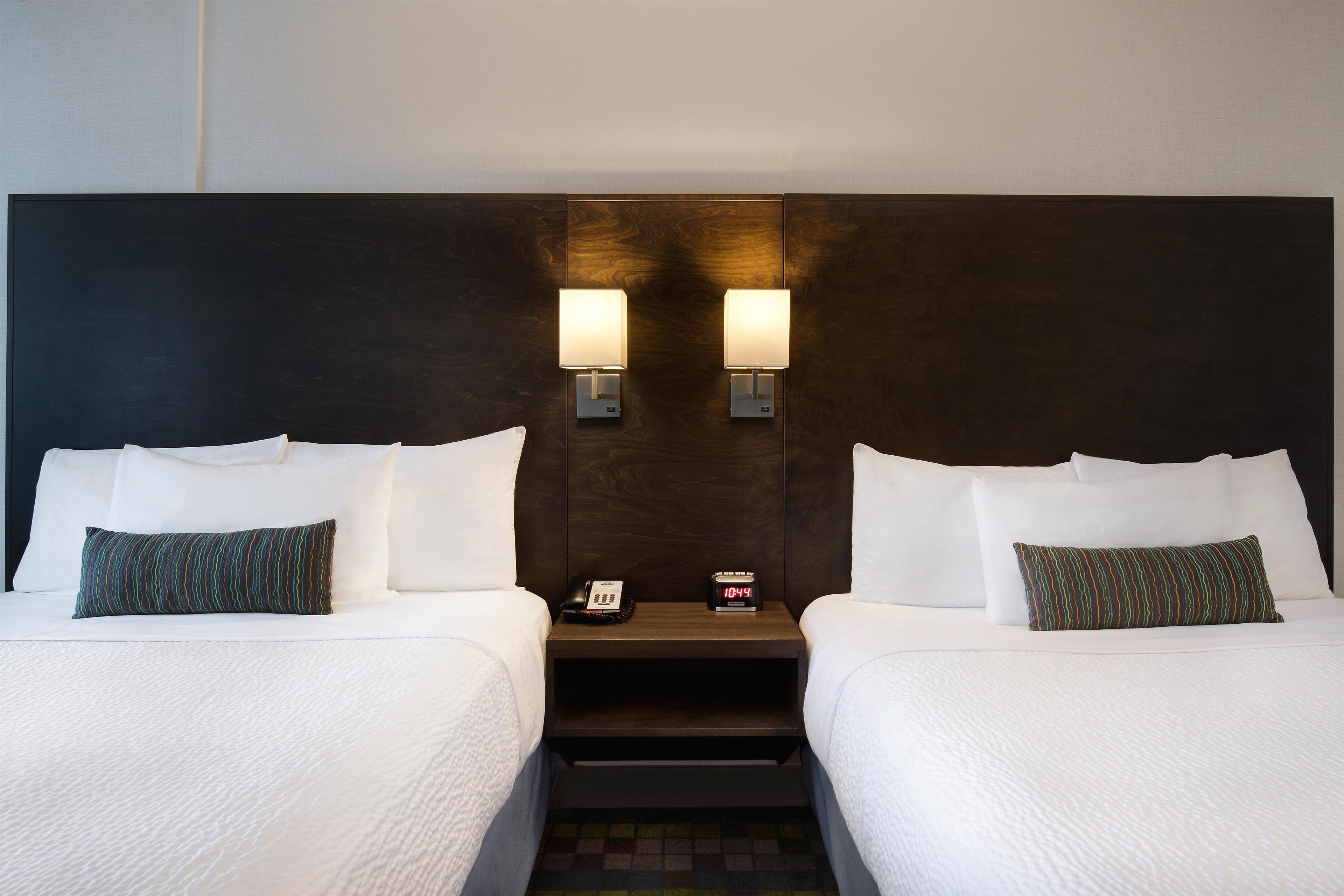 A hotel room with two beds, each with white bedding and patterned pillows, a nightstand with a phone and a digital clock, and wall-mounted lamps.