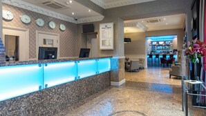 A hotel reception area with a marble counter, clocks on the wall, a bar in the background, and a seating area with a vase of flowers. - Sidney Hotel London Victoria  (London)