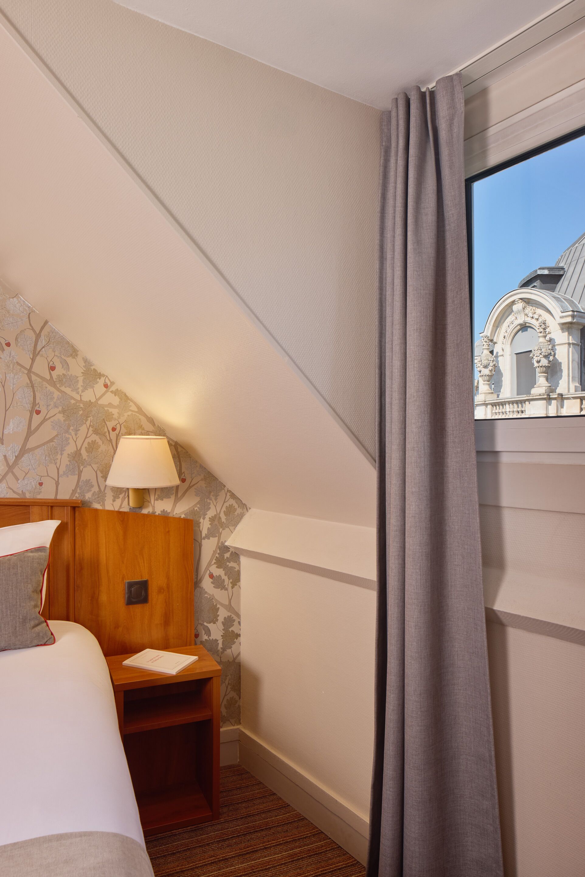 Una habitación de hotel con cama, mesita de noche, lámpara y ventana con cortinas.