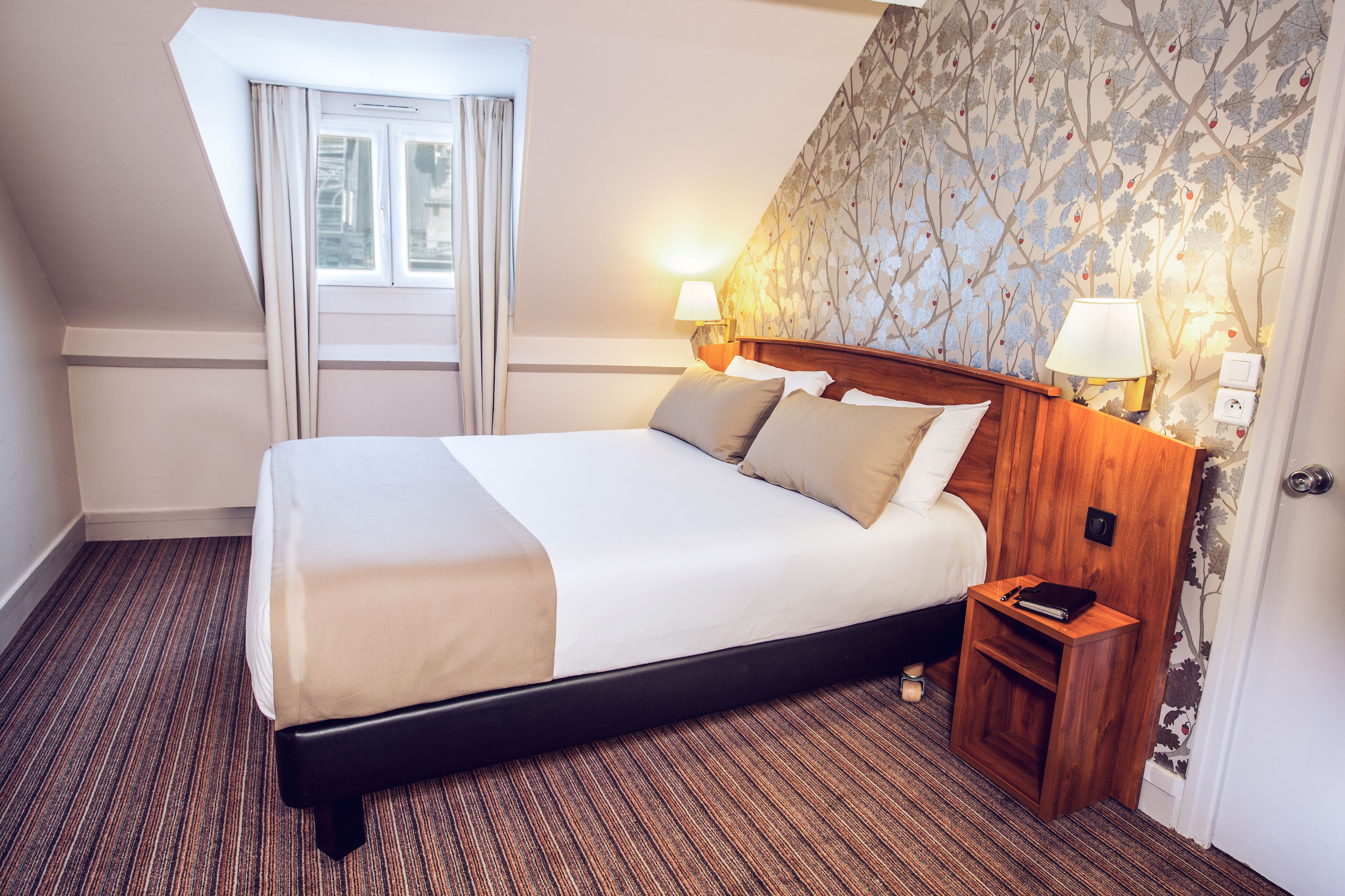 A hotel room with a large bed, two bedside lamps, a nightstand, and a window with curtains.