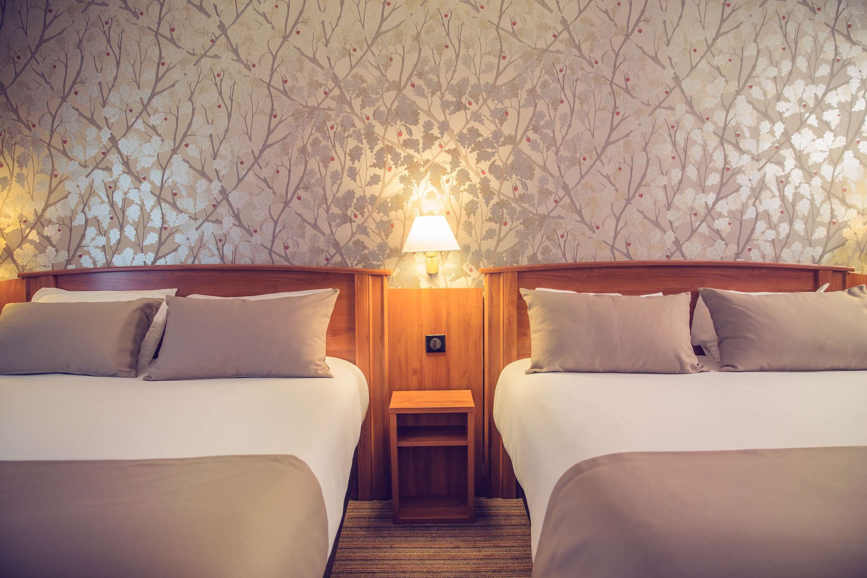 Een hotelkamer met twee bedden, een nachtkastje, een wandlamp en behang met een patroon.