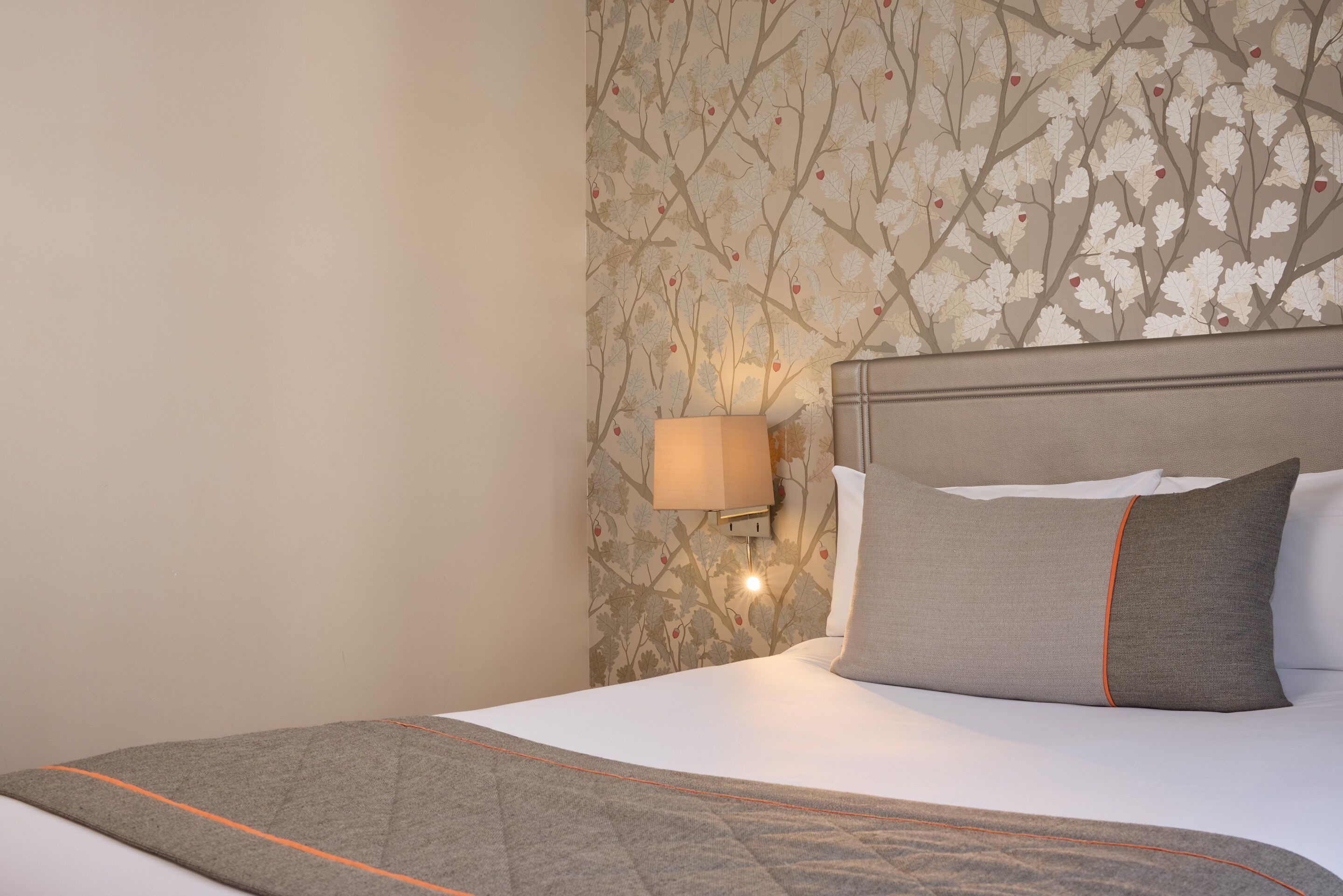 A neatly made bed with a grey blanket and pillows, a wall-mounted lamp, and a floral wallpaper pattern.