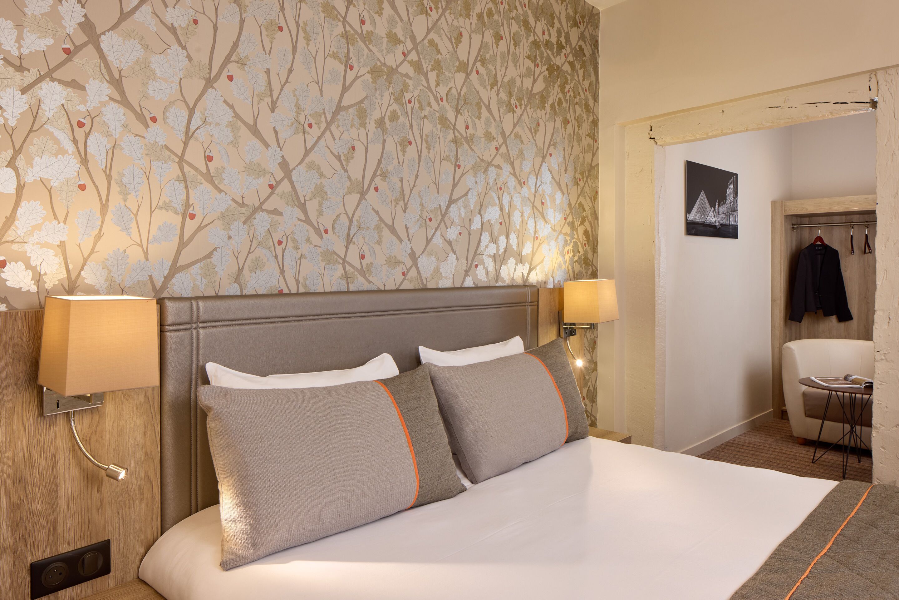 Een net opgemaakt bed met twee kussens, een hoofdbord, en een wand met een bloemenpatroon. Er staat een lamp aan de muur en een kapstok met een jas.