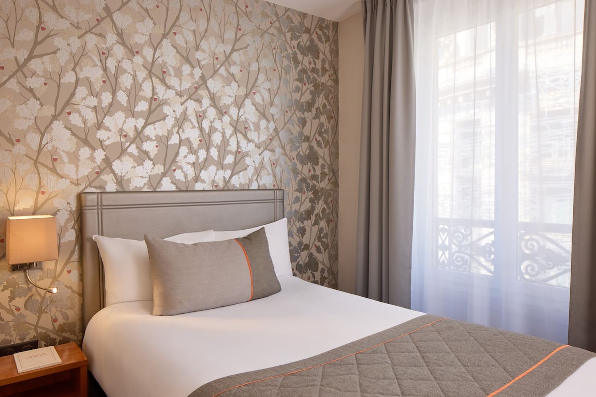 Hotellihuone, jossa on sänky, yöpöytä, lamppu ja ikkuna, jossa on verhot.