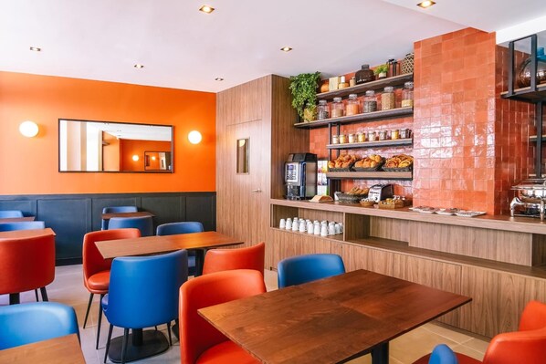 Un restaurante con paredes anaranjadas, mesas de madera y sillas azules. Se observan estantes con frascos y una máquina de café.