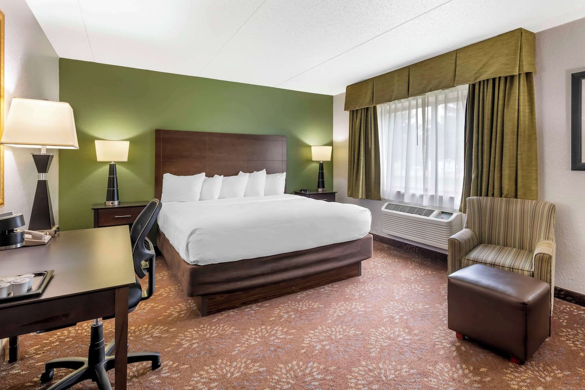 A hotel room with a large bed, a desk, a chair, a small ottoman, and a window with curtains.