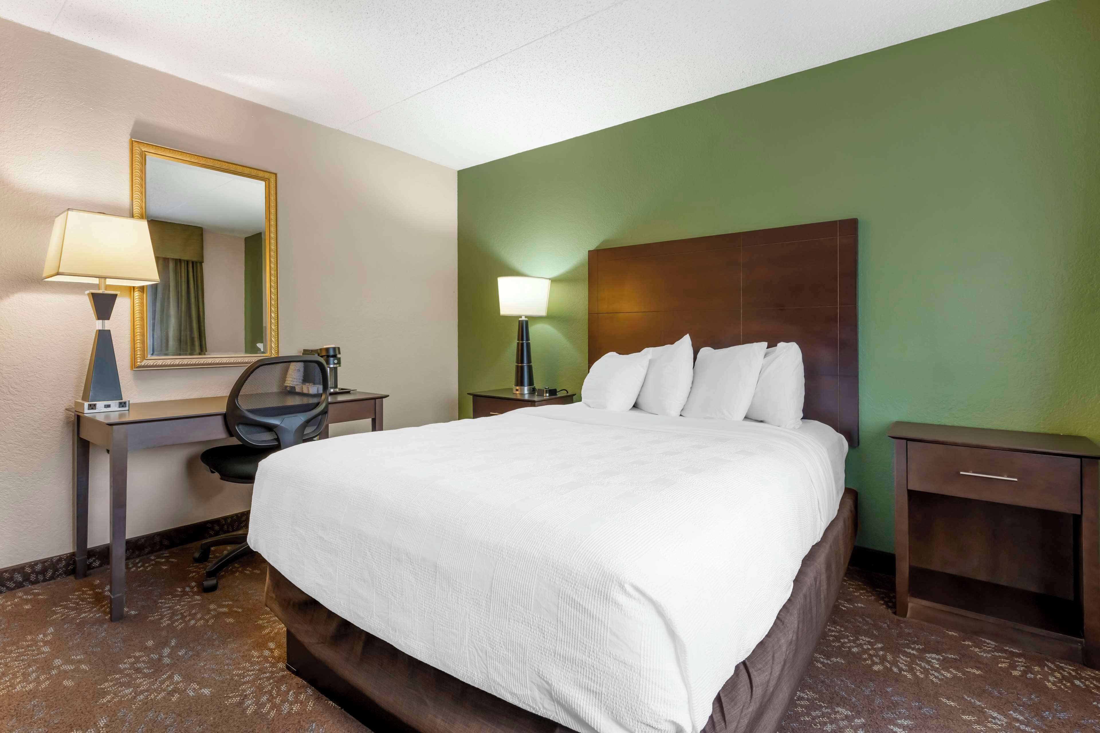 A hotel room with a large bed, a desk with a chair, a nightstand with a lamp, and a mirror on the wall.