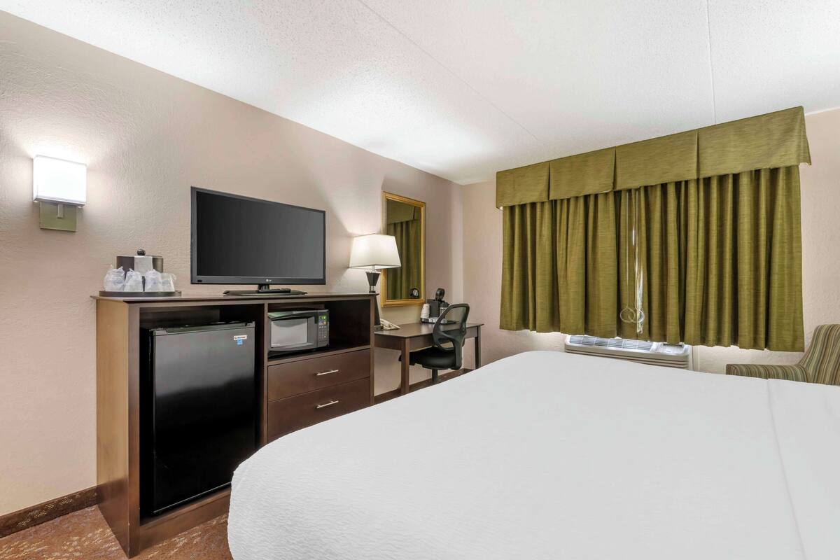 A hotel room with a bed, a desk with a computer, a television, a refrigerator, and a window with curtains.