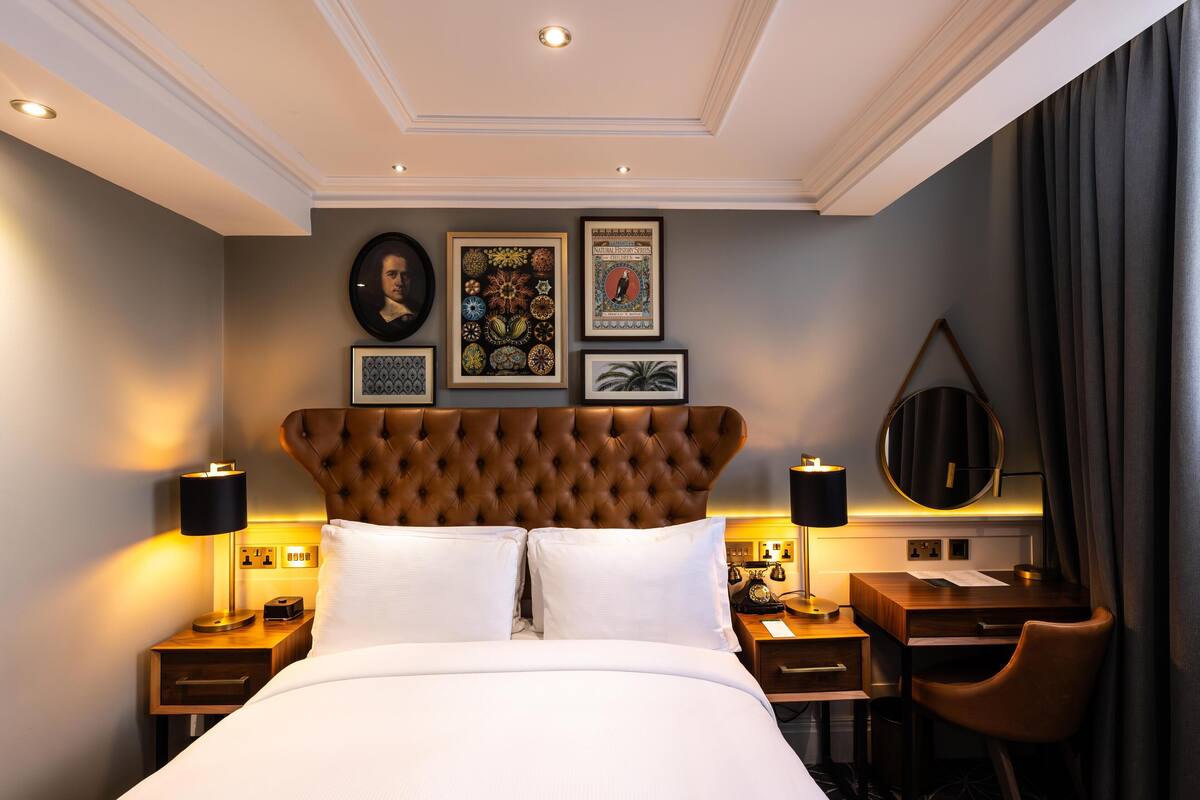 A hotel room with a tufted headboard, a bed with white linens, a desk with a chair, a round mirror, and framed artwork on the wall.