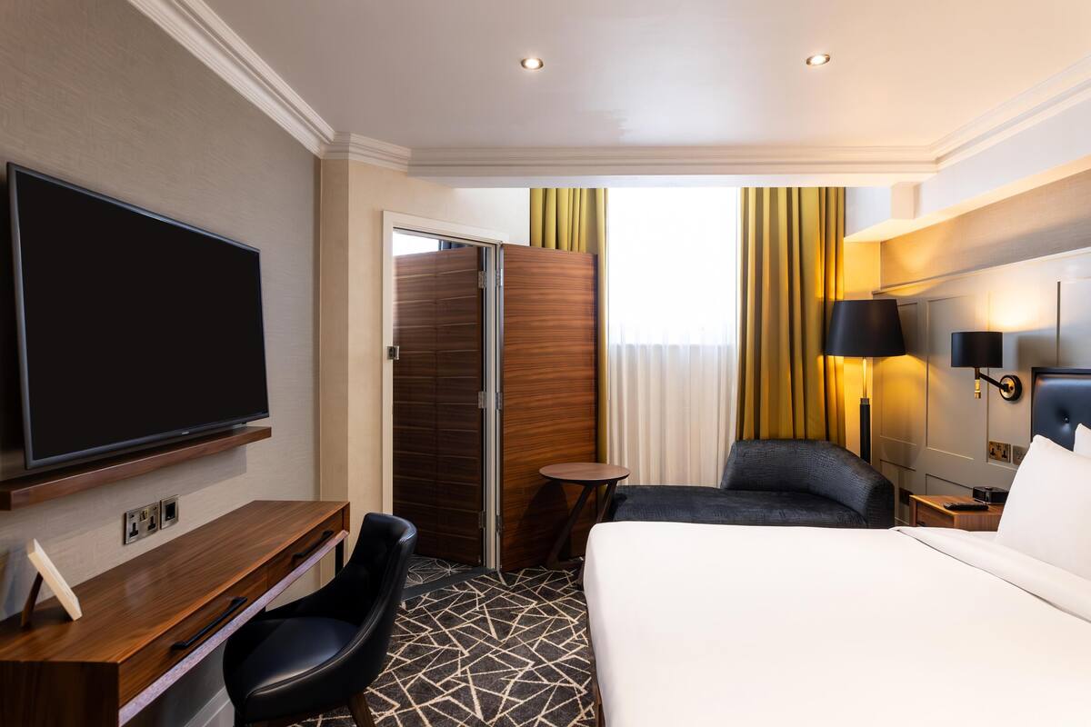 A hotel room with a bed, a desk with a flat-screen TV, a chair, a small table, and a lamp.