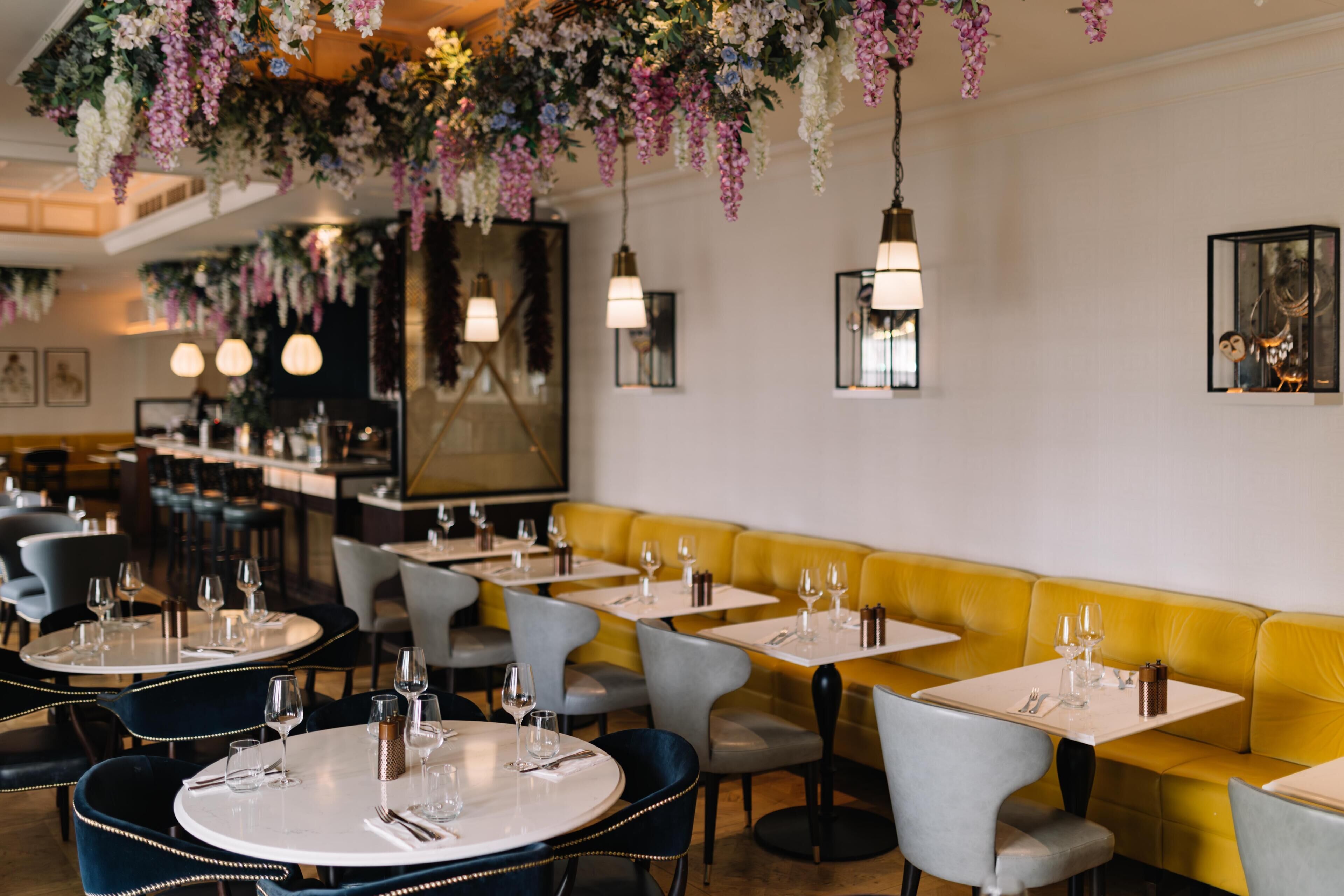 Interior de restaurante con mesas redondas, sillas grises, banquetas amarillas, adornos florales colgantes y lámparas fijadas a la pared.