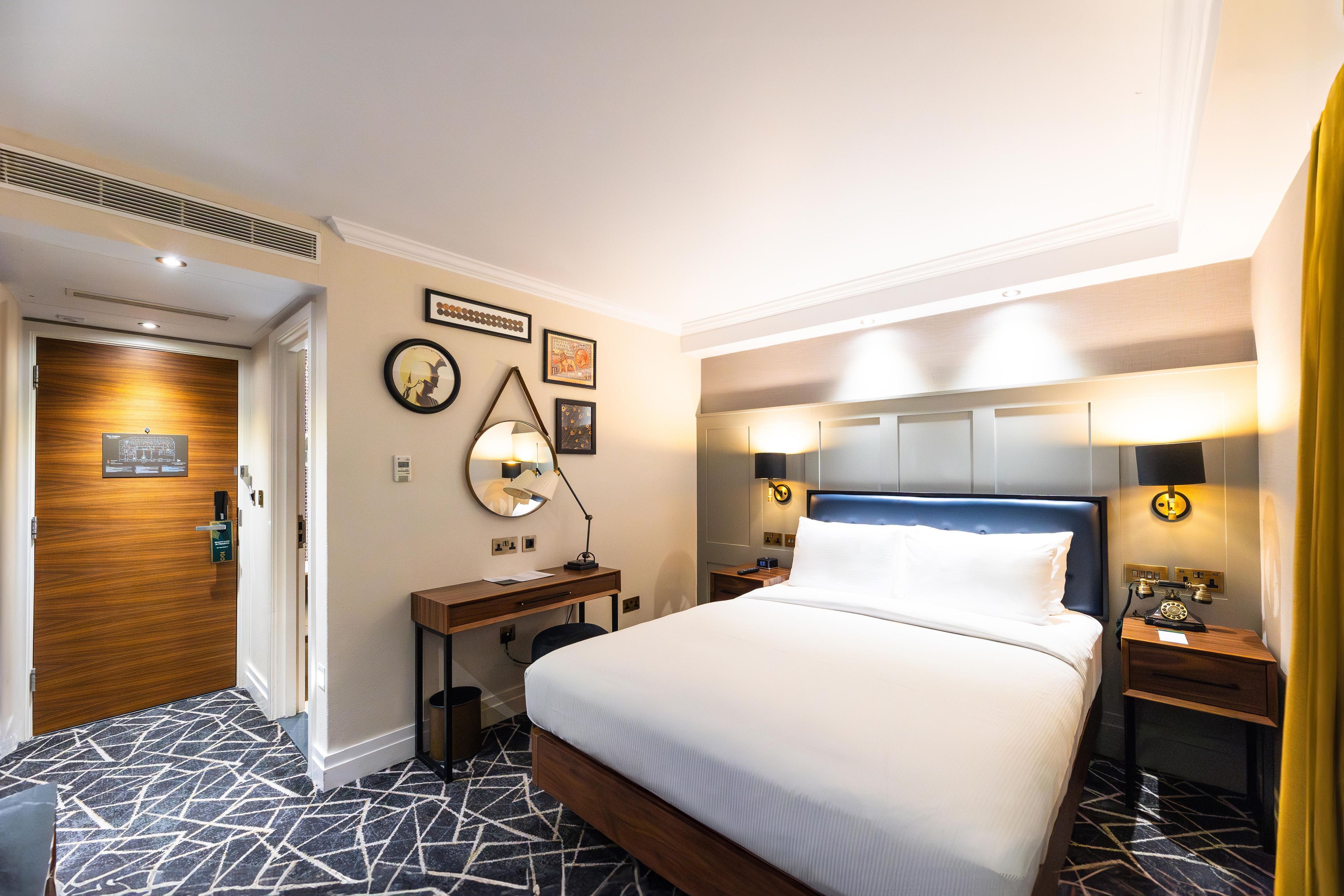 A hotel room with a bed, bedside tables, a desk, and a mirror. There are wall-mounted lamps and a clock on the wall.