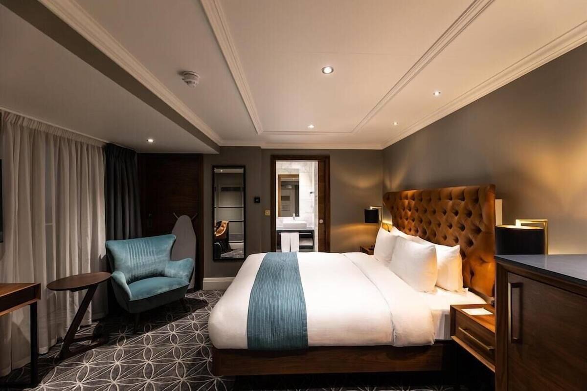 A hotel room with a large bed, a sitting area with a chair, a bedside table, and a wardrobe.