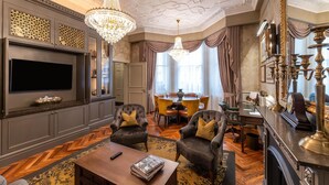 A luxurious living room with a large flat-screen TV, two armchairs with cushions, a wooden coffee table, a chandelier, and a dining area with a table and chairs. - 100 Queen's Gate Hotel London Kensington, Curio Collection by Hilton (London)